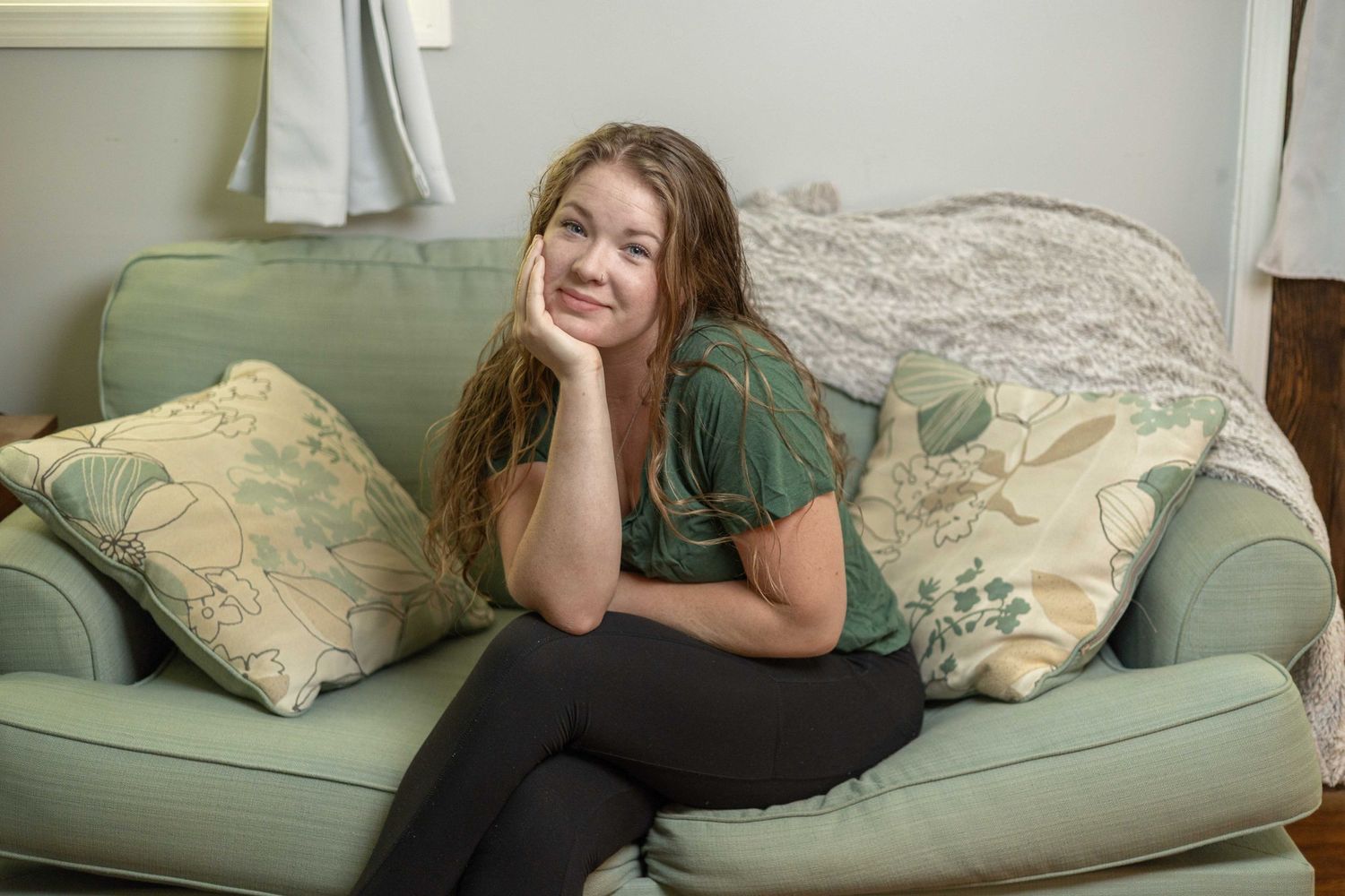 Someone relaxes casually on a sage green couch with decorative floral throw pillows in a bright interior setting.