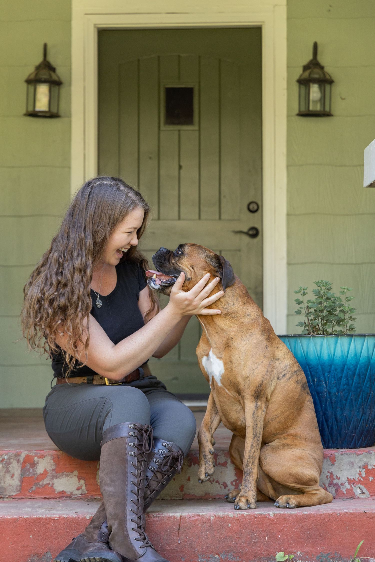 Person playing with brown dog on front steps of green house with hanging lanterns.