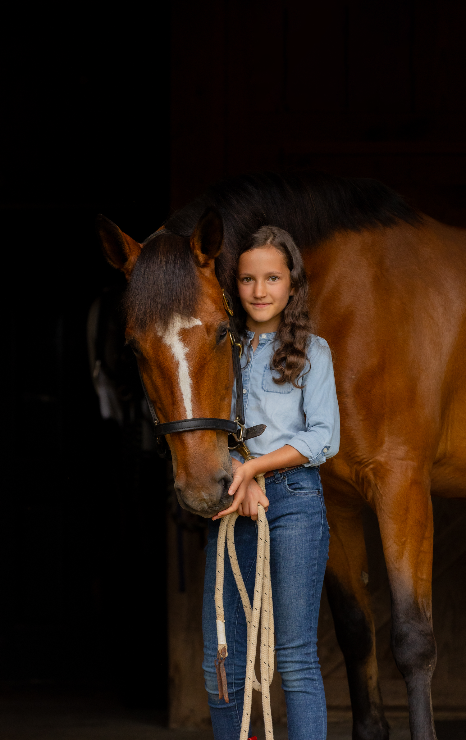 Young equestrian stands with a bay horse in a dark barn wearing denim and a chambray shirt.