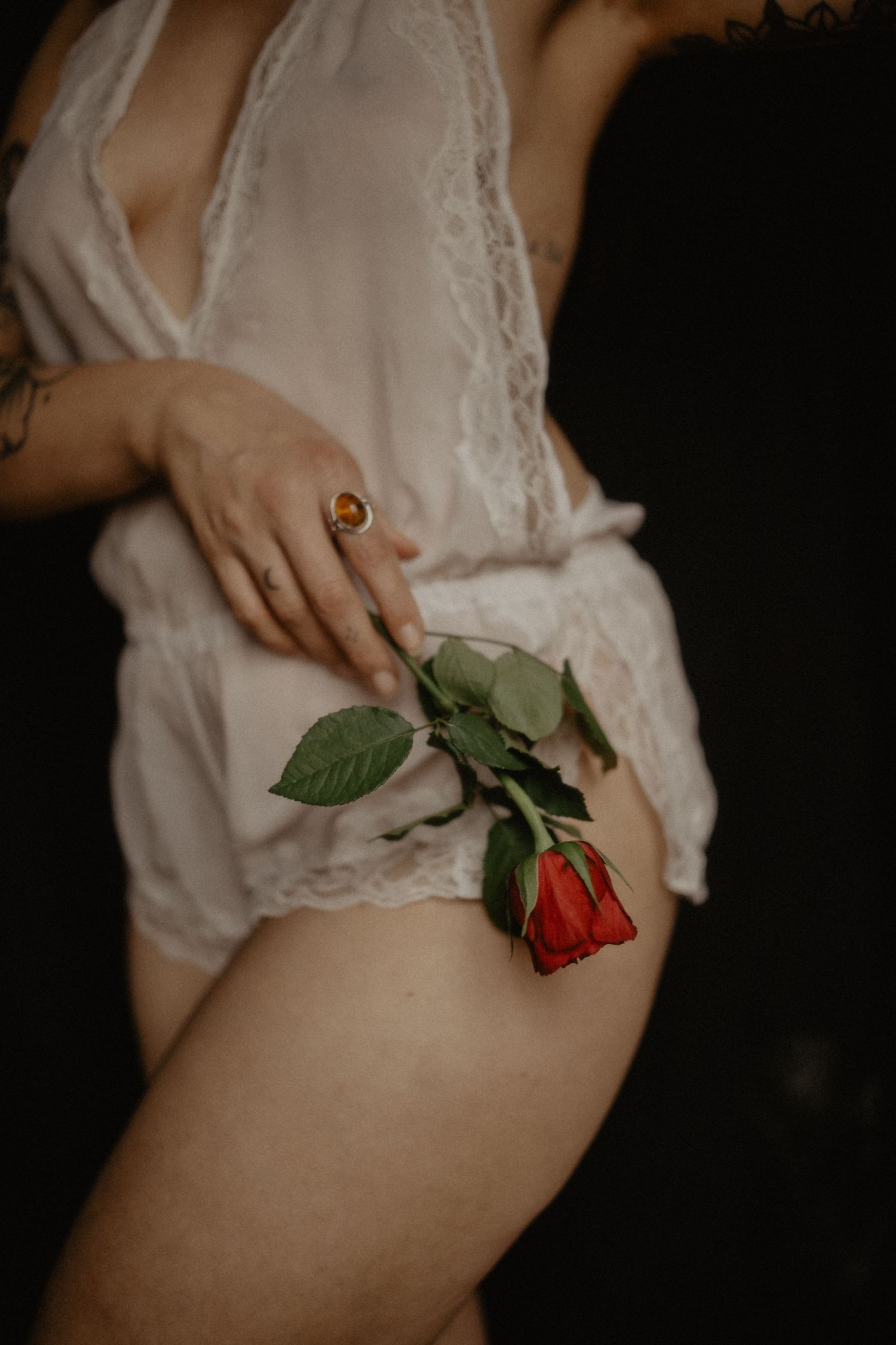 A delicate hand holding a red rose against white lace lingerie in a moody boudoir setting.