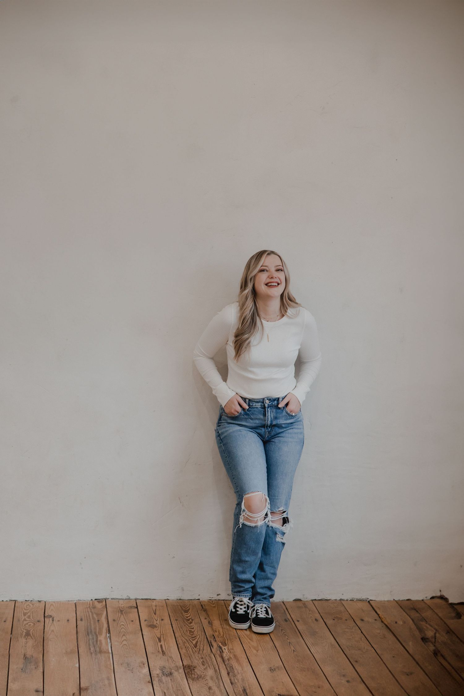 A casual lifestyle portrait of someone wearing a white sweater and ripped jeans against a beige wall.