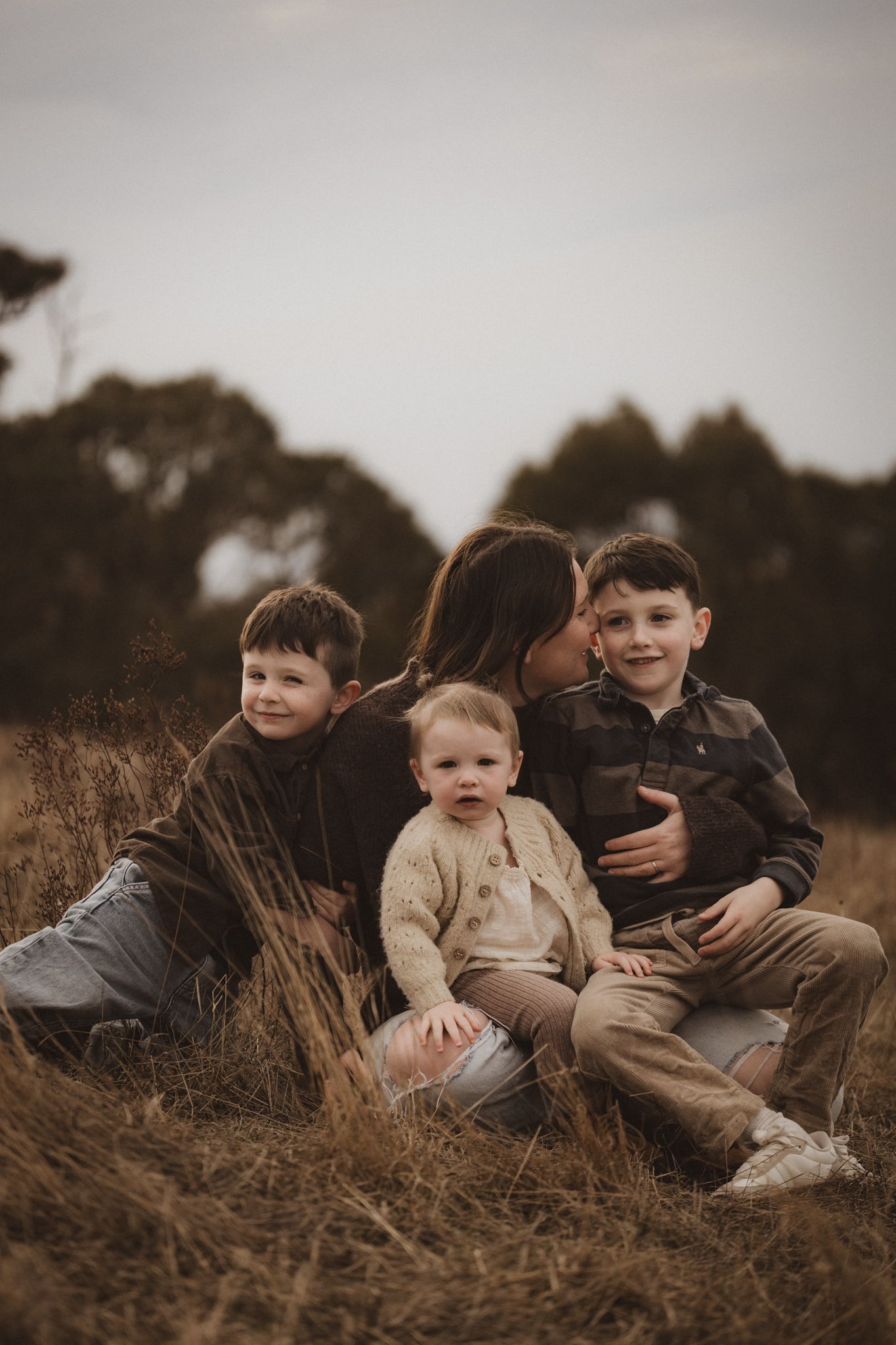 Moody family portrait in tall grass field with children sitting together at dusk with trees in background.