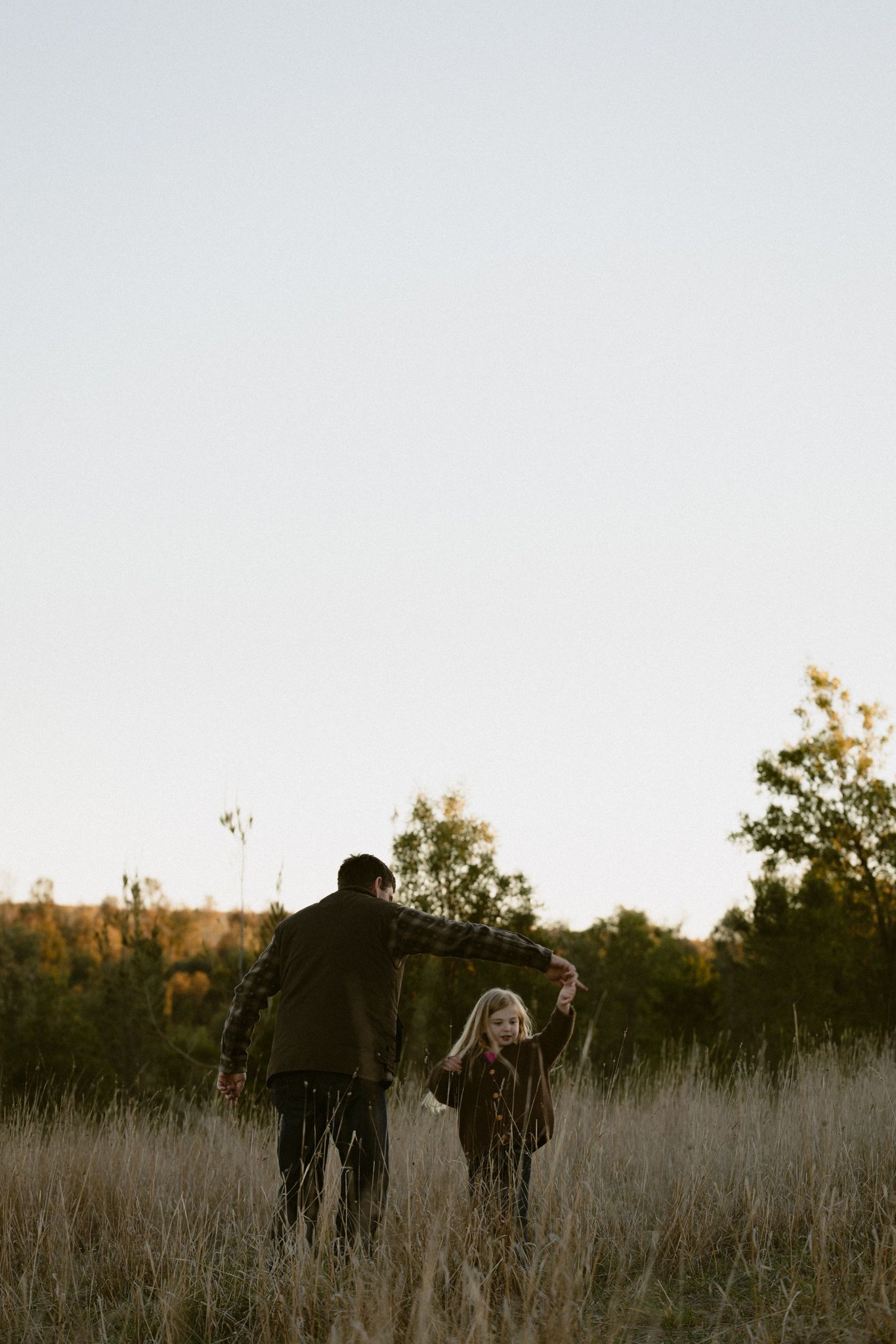Two figures stand together in a golden grassy field at sunset with trees silhouetted in the background.