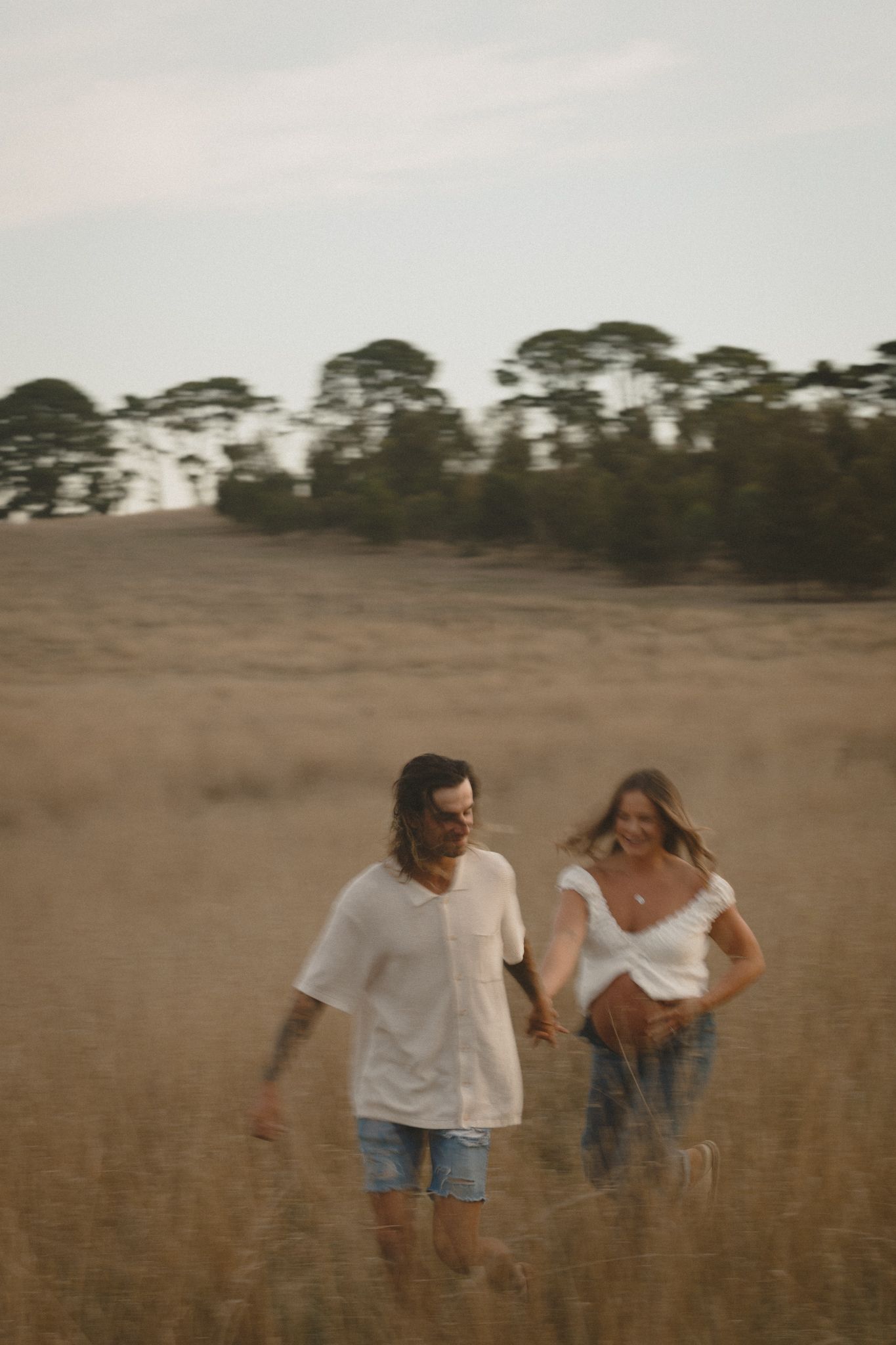 Two people walk through a golden field at sunset with tall pine trees silhouetted against the sky in the background.