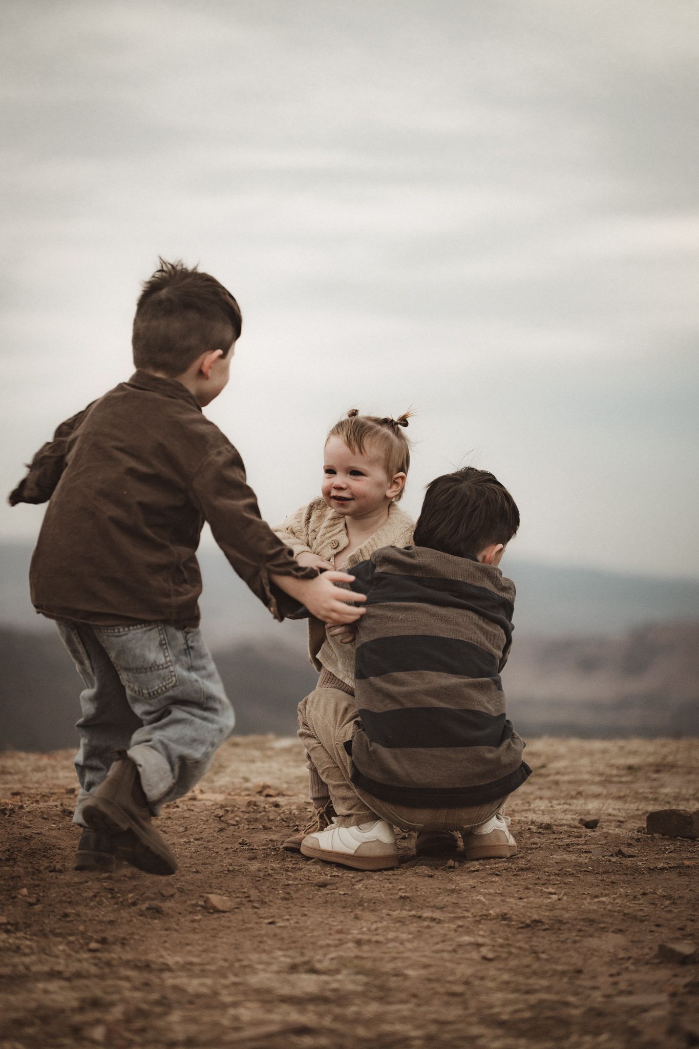Three children play together outdoors on a hilltop against an overcast sky in muted brown tones.