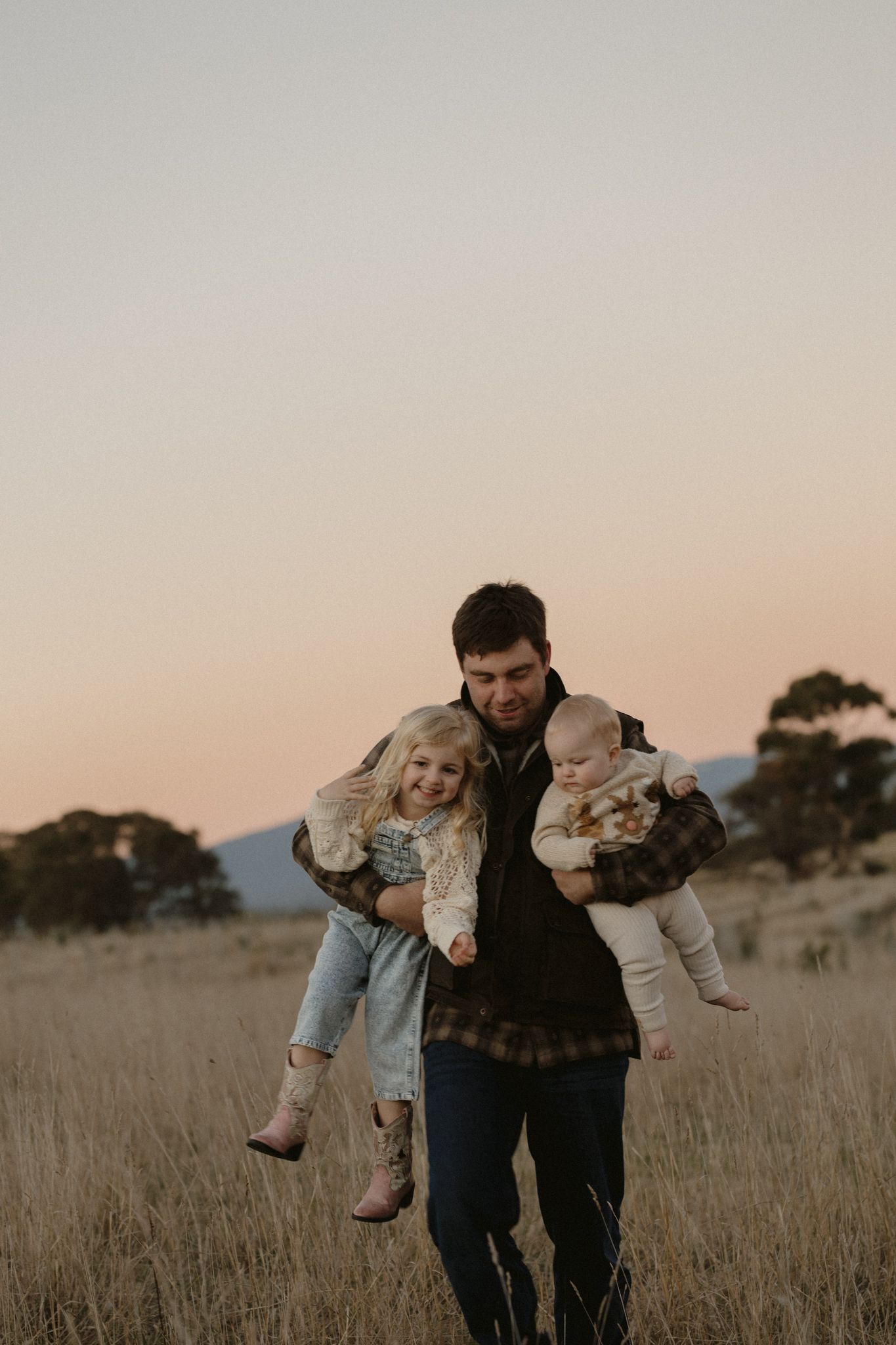 A person carries two children through a rustic field at sunset with mountains in the background.