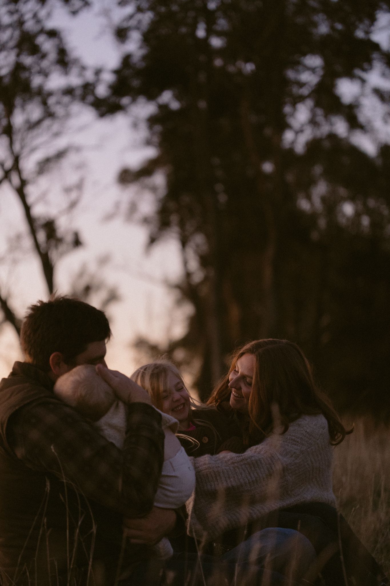 Silhouettes of people embracing outdoors against a dusky sky with trees in the background during sunset.