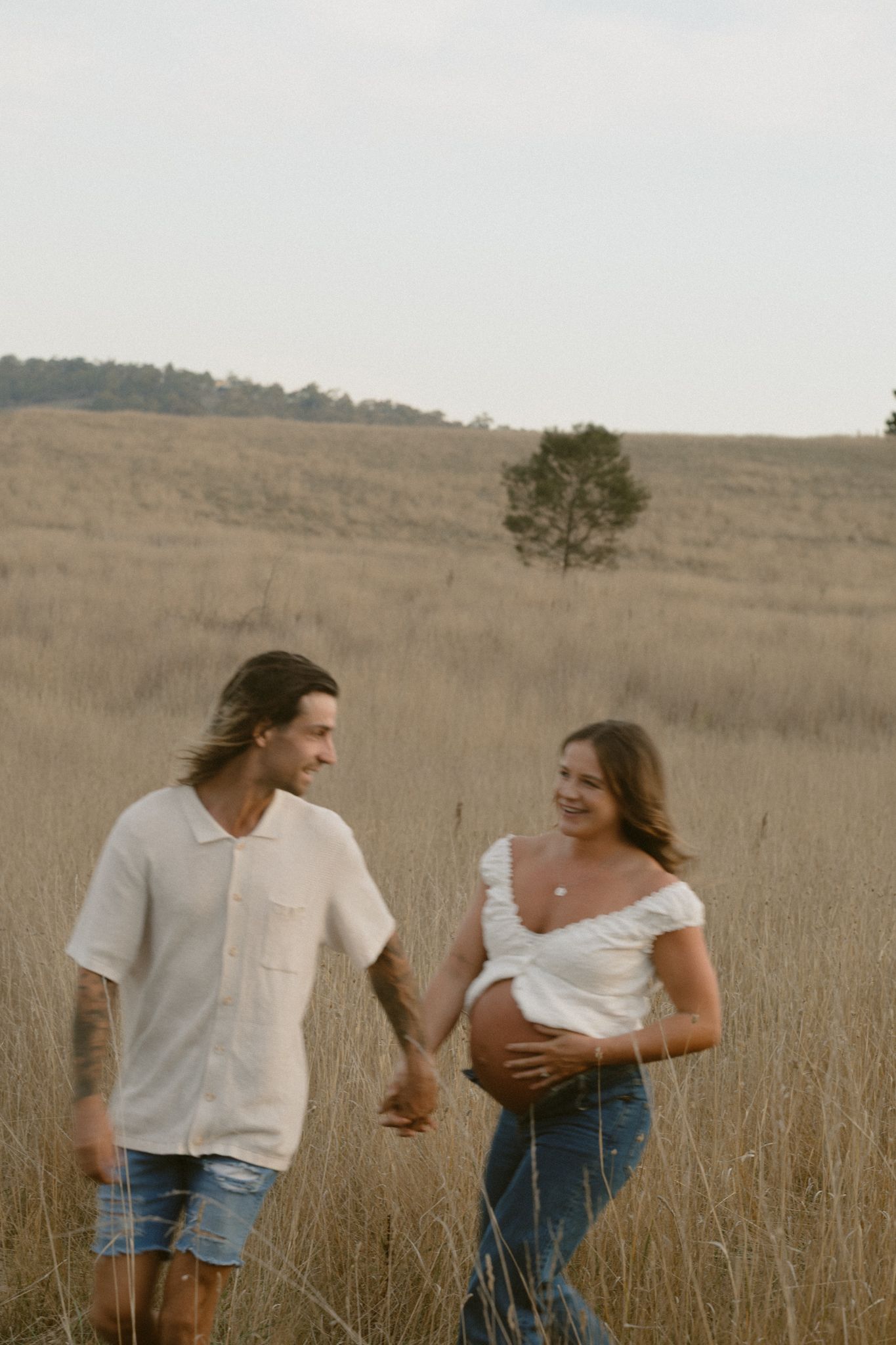 Couple holding hands in a tall grass field during golden hour with single tree in background.