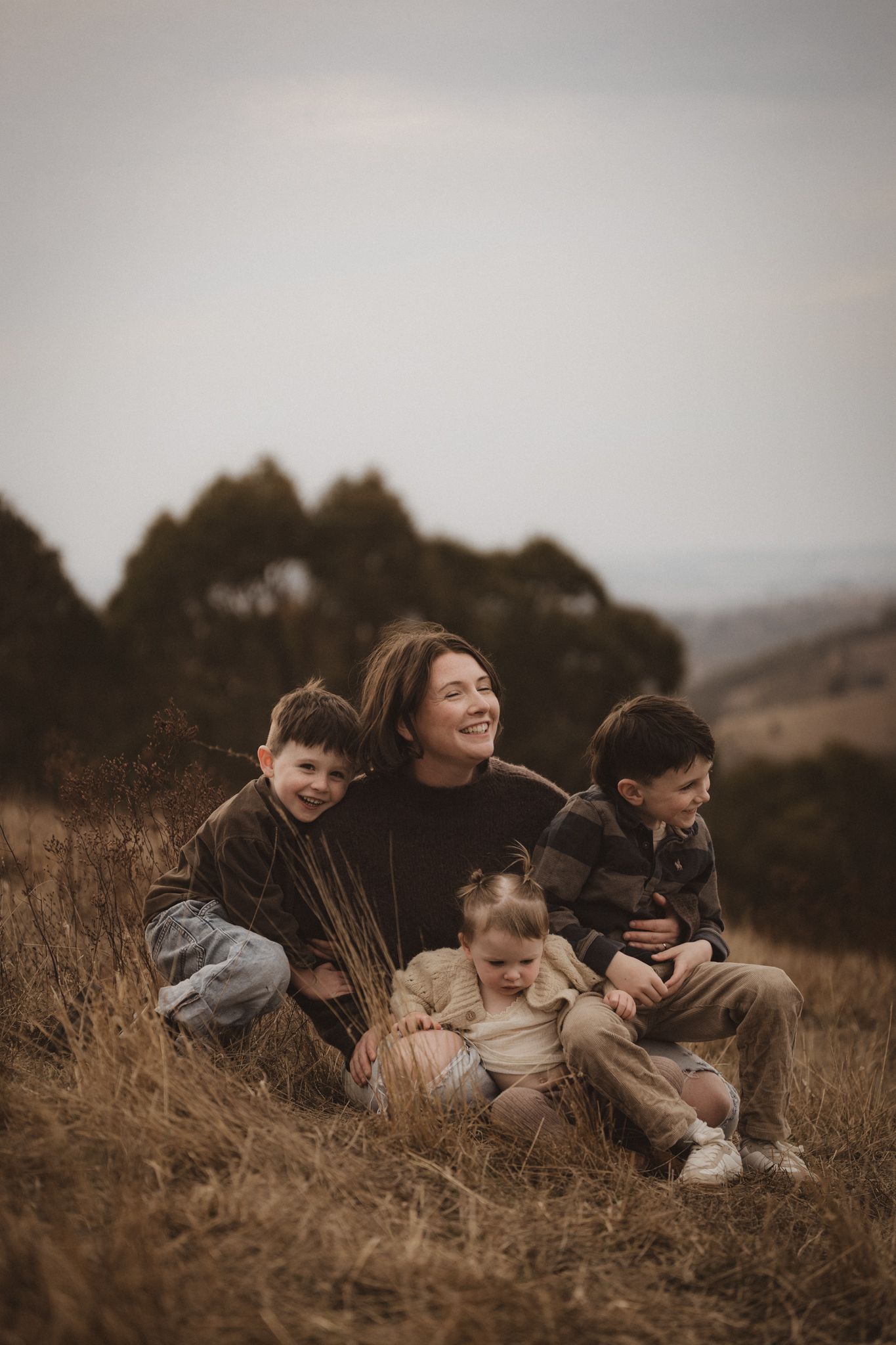 A family sits together in golden grass on a hillside during sunset, creating a warm and intimate outdoor portrait.