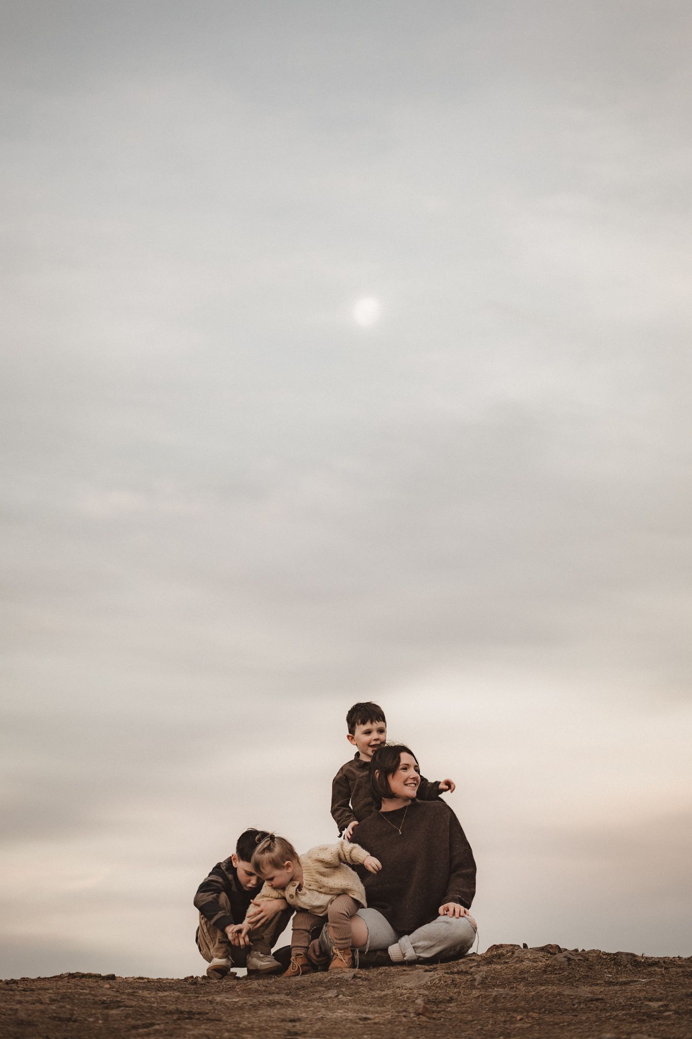 Family sits together on hilltop against moody cloudy sky with dim sun peeking through at sunset.