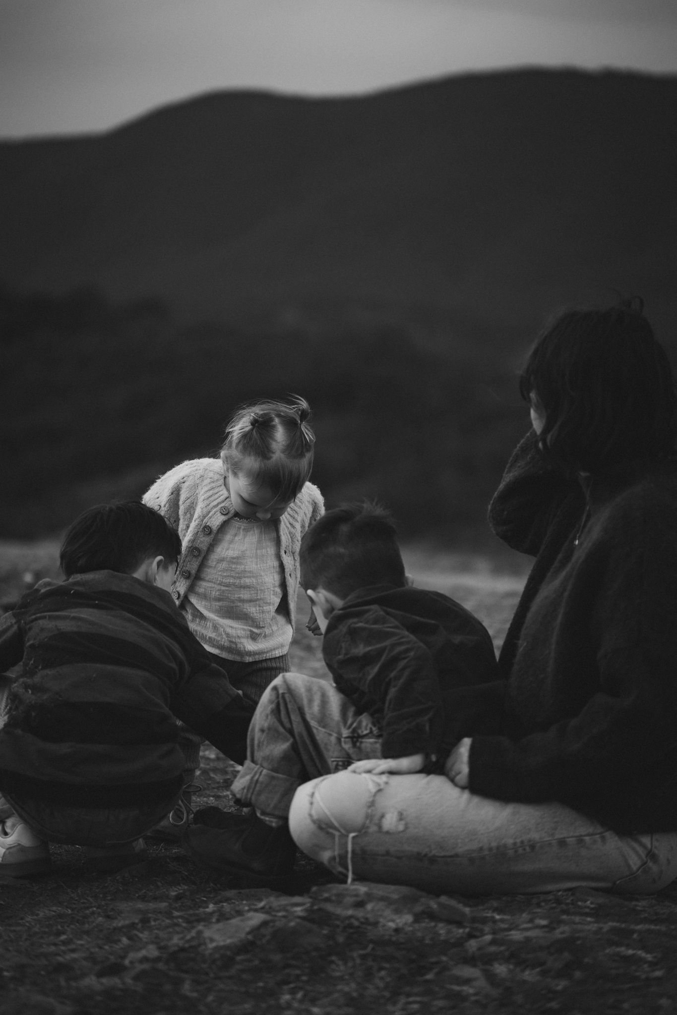 A black and white image of people sitting together on a hillside at dusk, creating a moody silhouette against mountains.