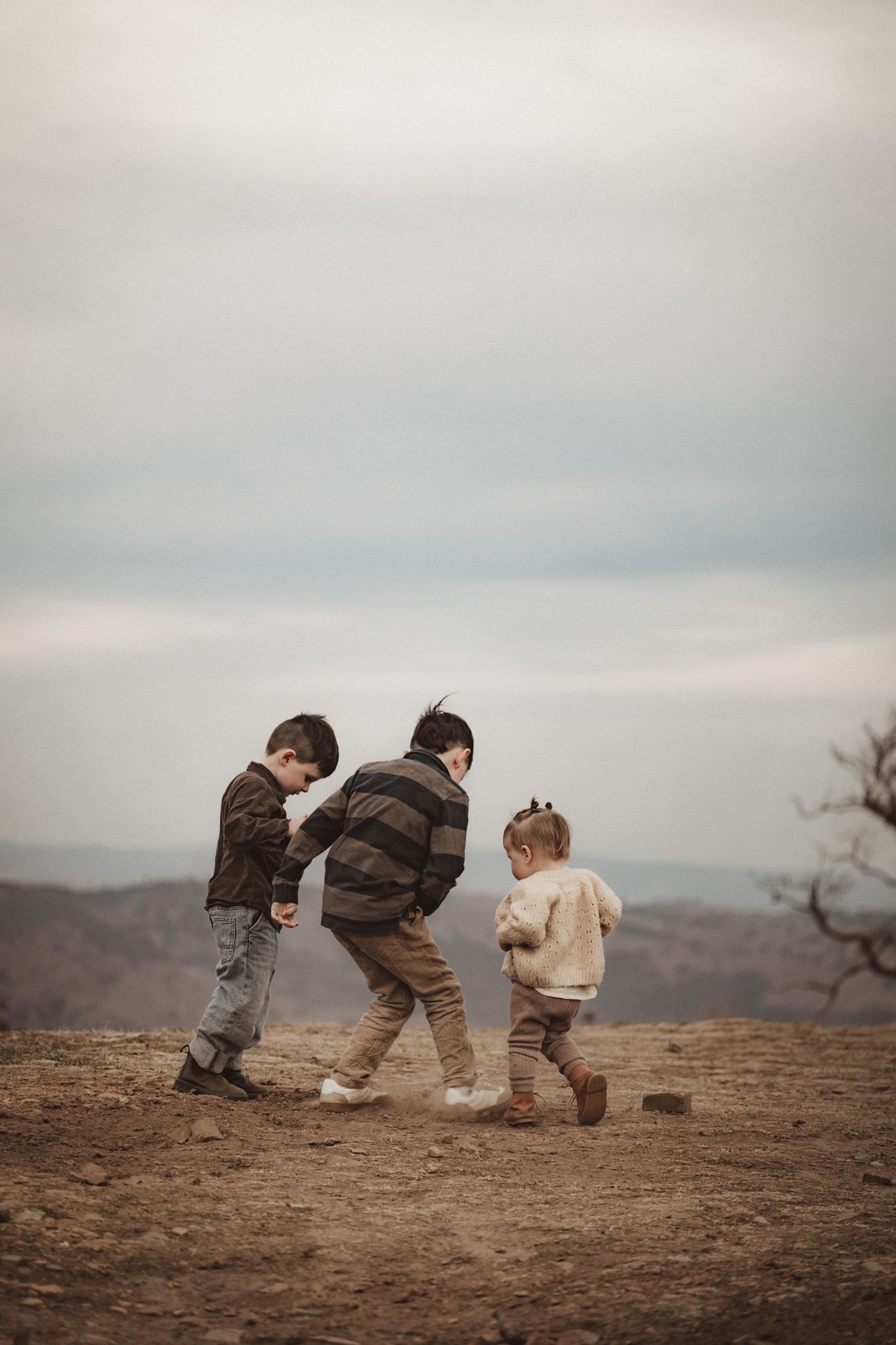 Three children in warm clothes playing together on a hilltop against a cloudy sky at sunset.