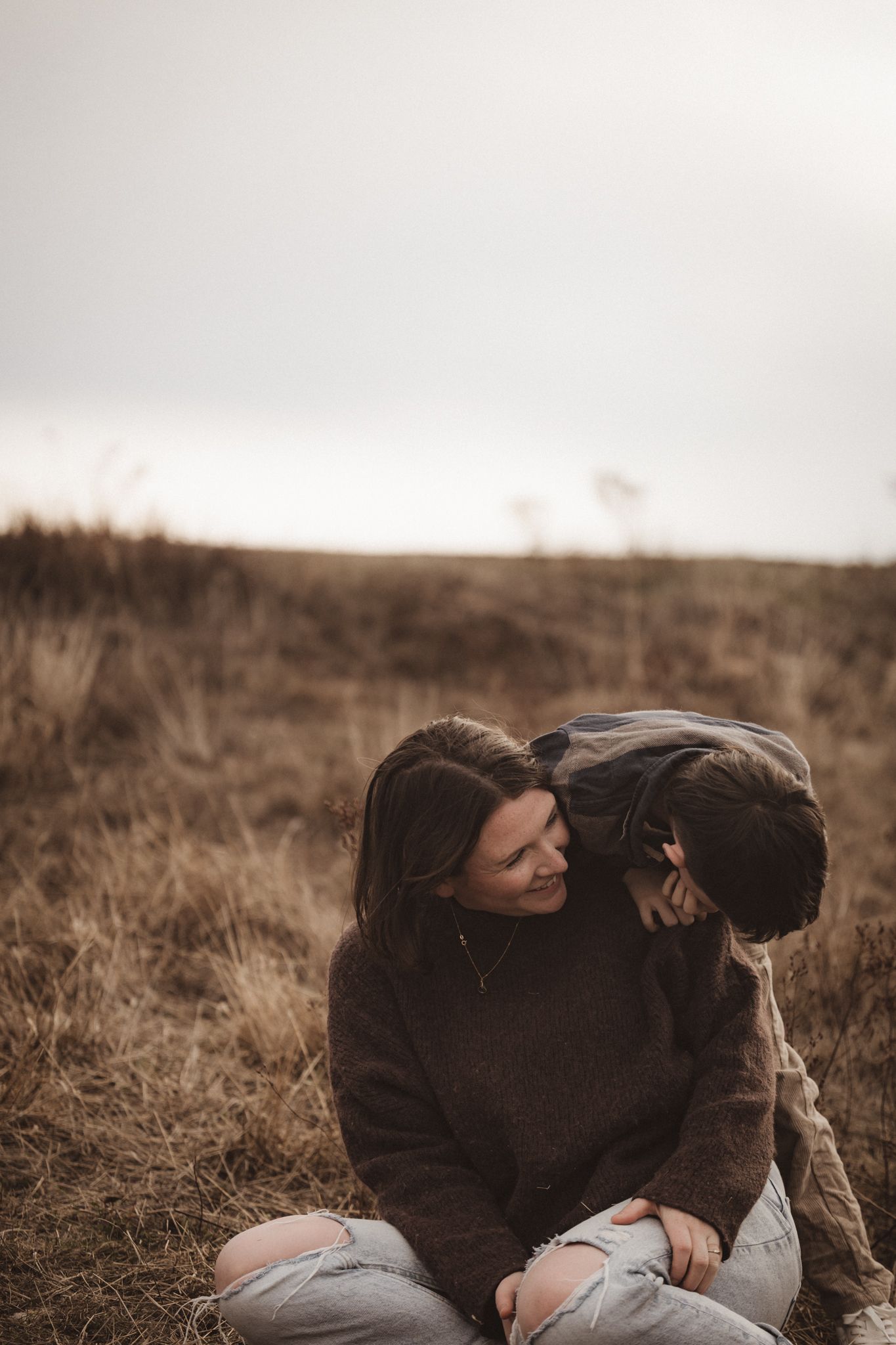 Person in a dark sweater sits in a brown grassy field on an overcast day enjoying a quiet moment outdoors.
