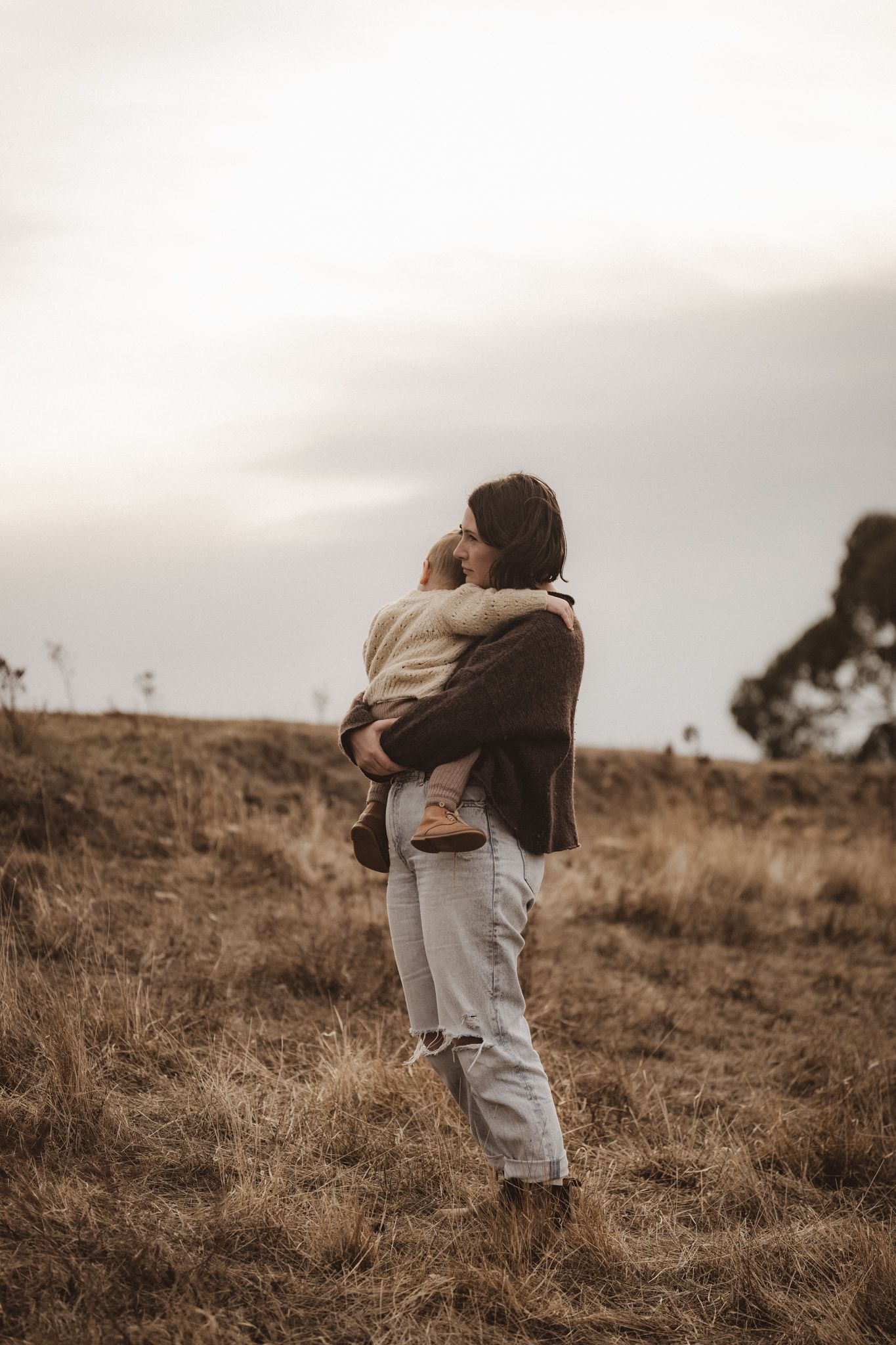 Figure stands in tall brown grass on hillside holding child during moody overcast evening.