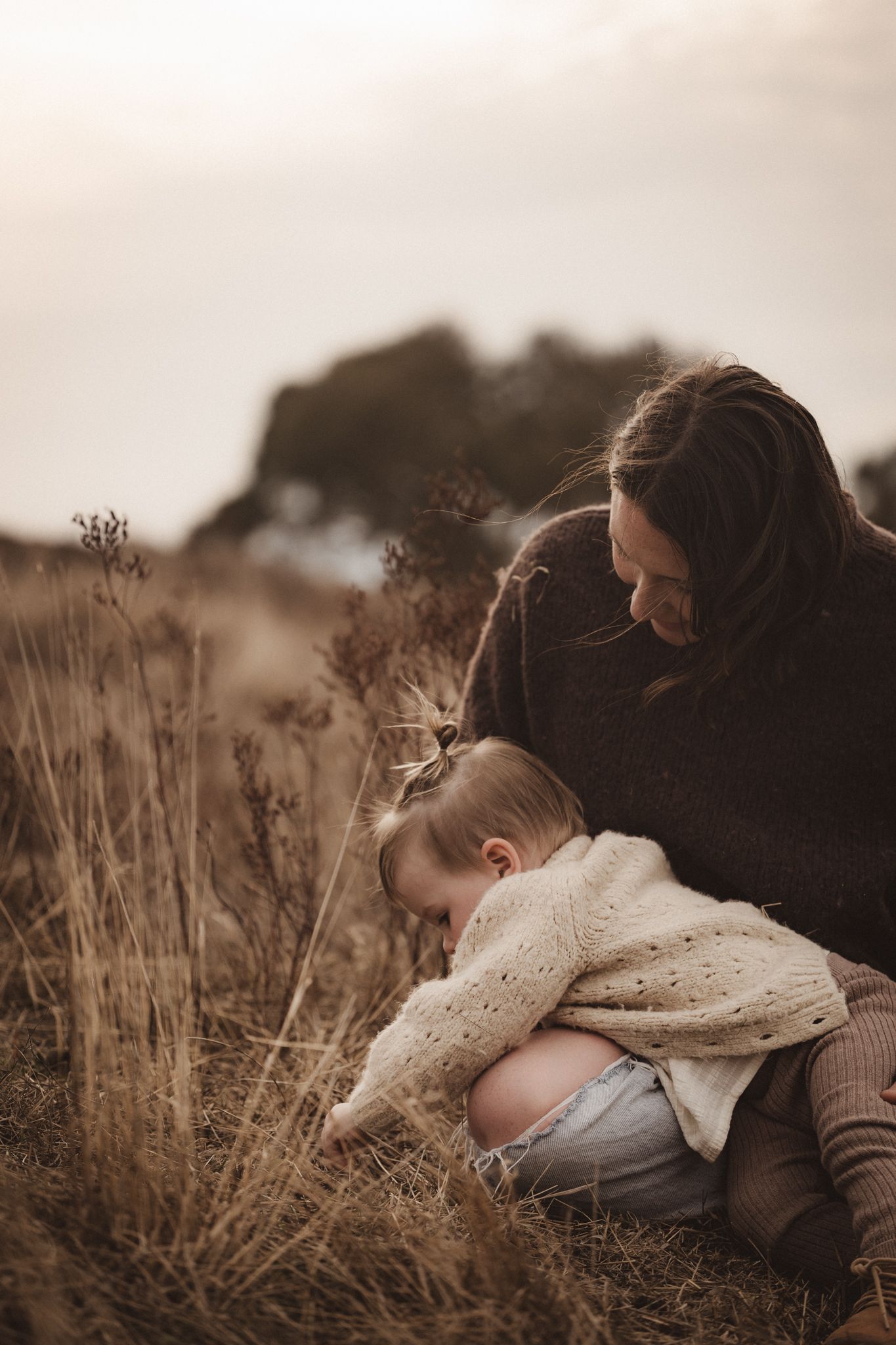 A tender moment between parent and child exploring dry grass together in warm sunset lighting.