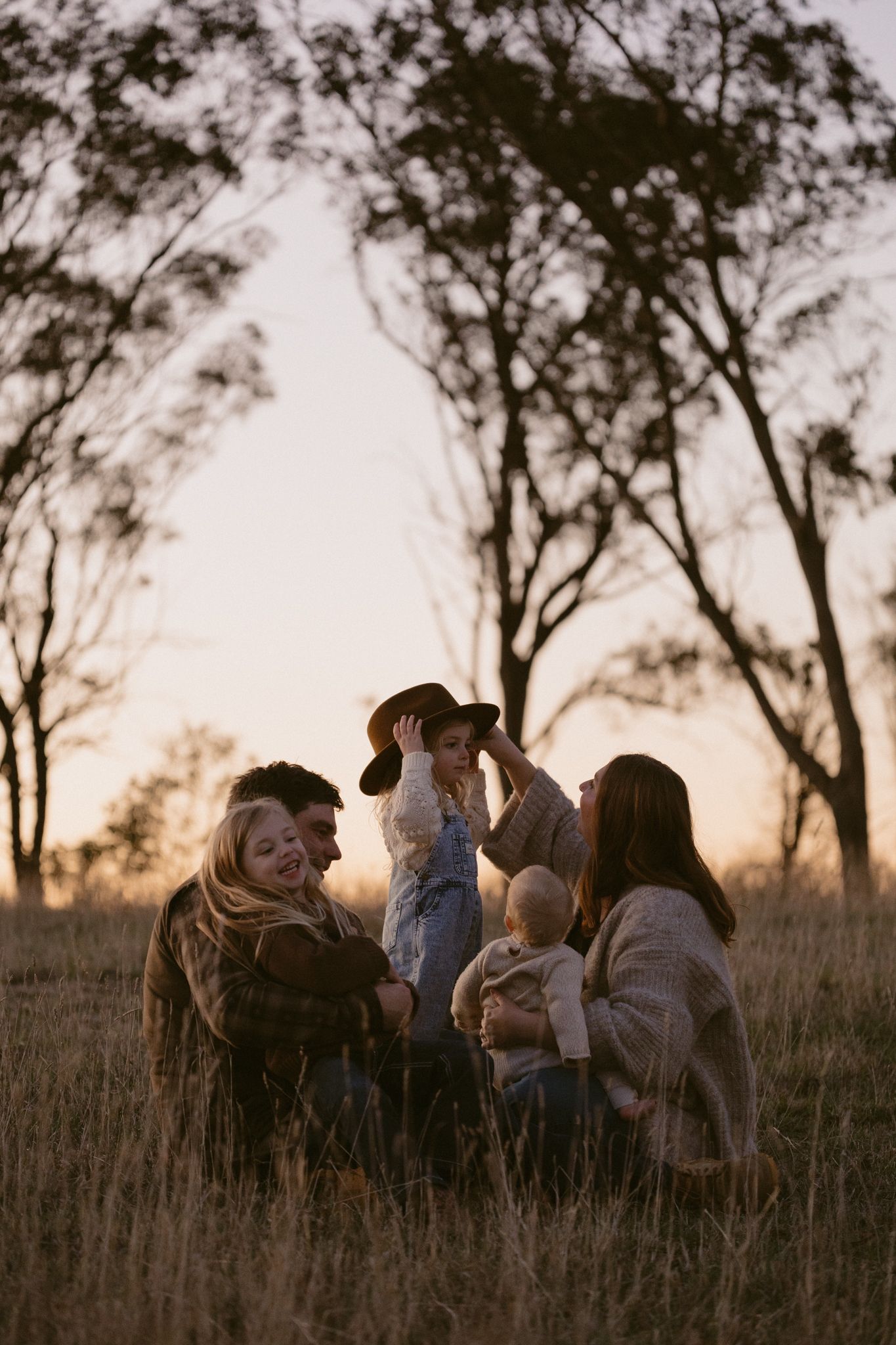 A group sits together in a grassy field at sunset as silhouetted trees frame the peaceful outdoor scene.