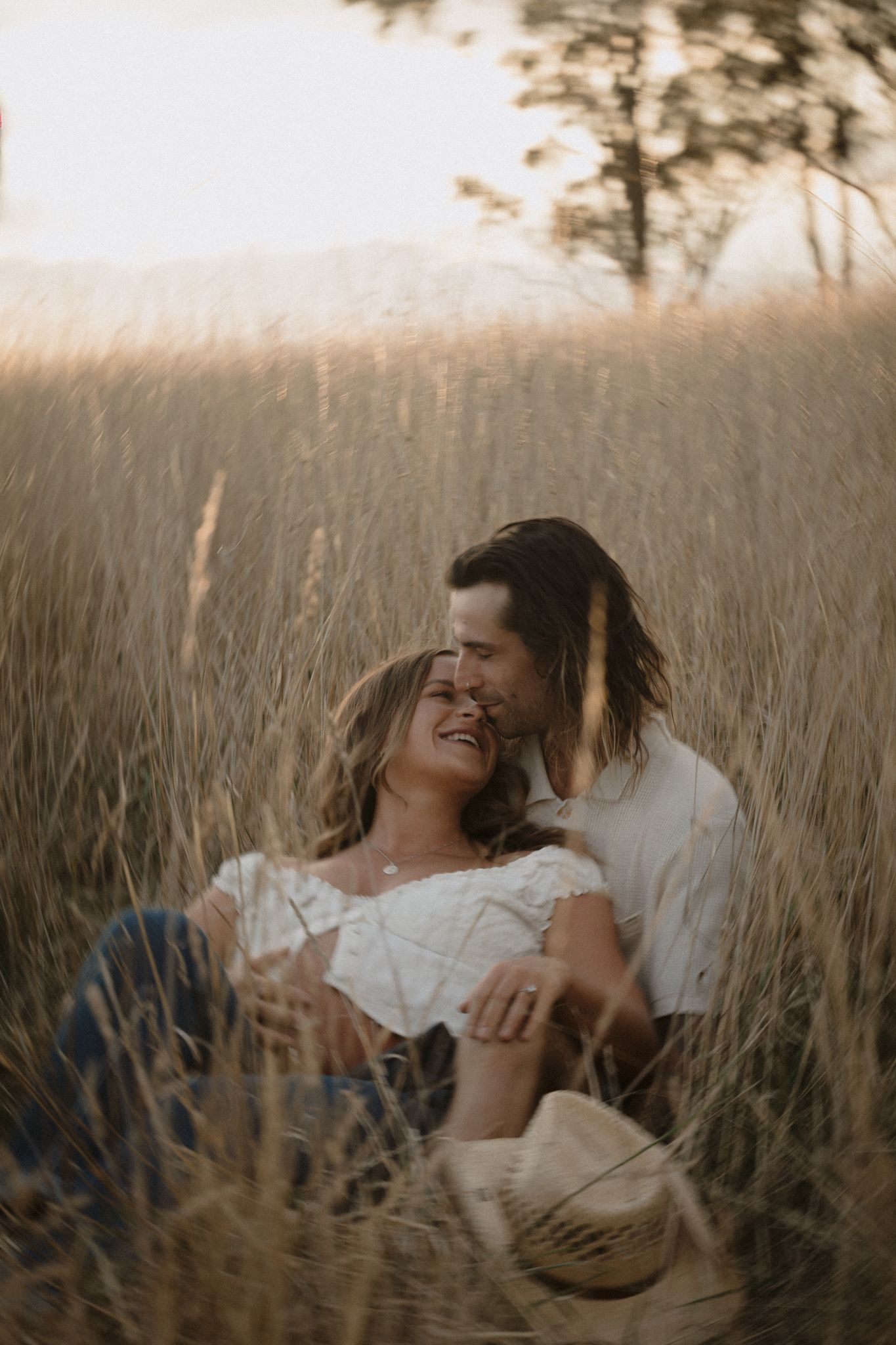 A romantic couple laughing together while sitting in tall grass during sunset.