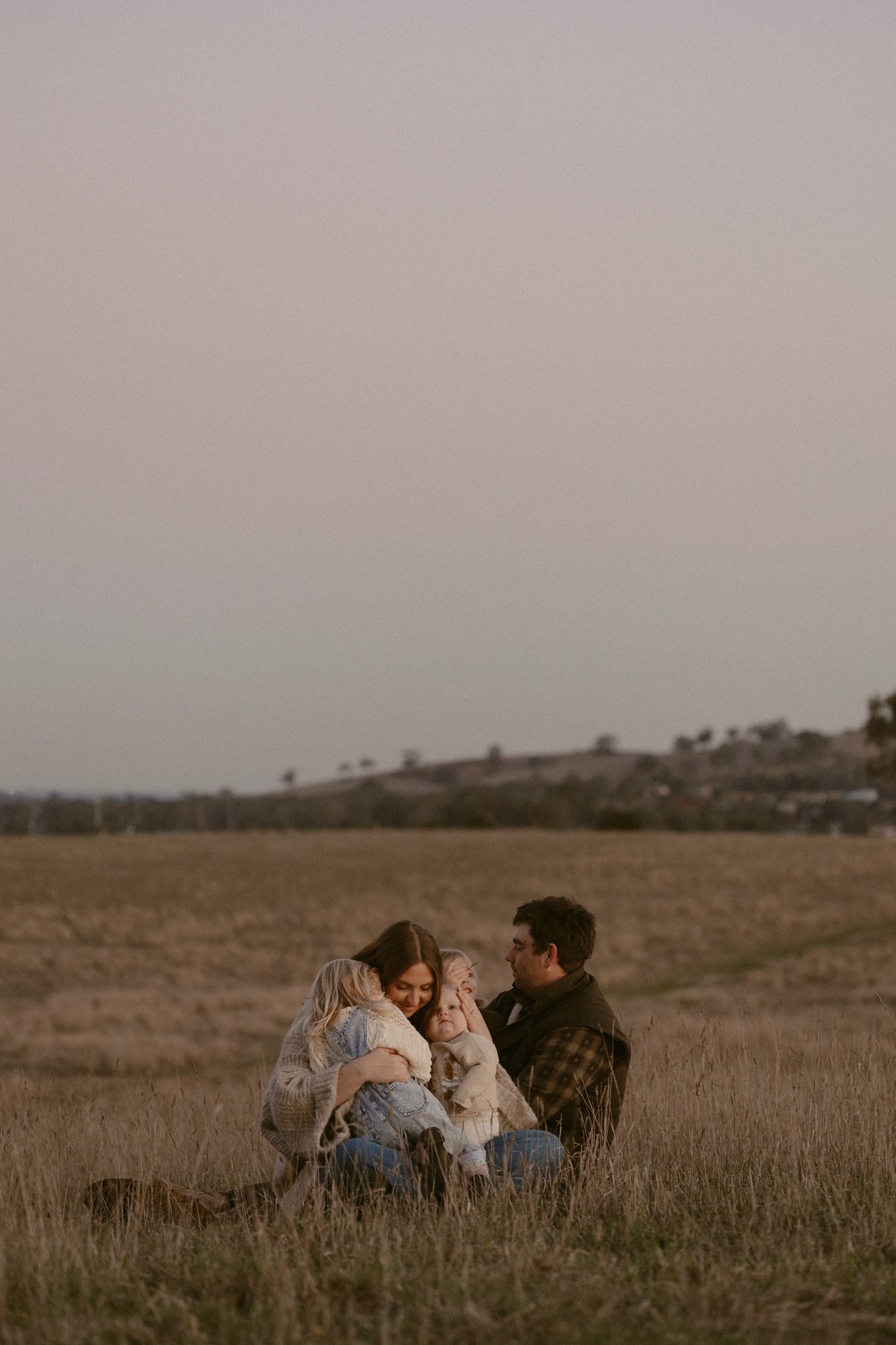 Three people sit together intimately in a grassy field at golden hour, creating a warm and atmospheric scene.