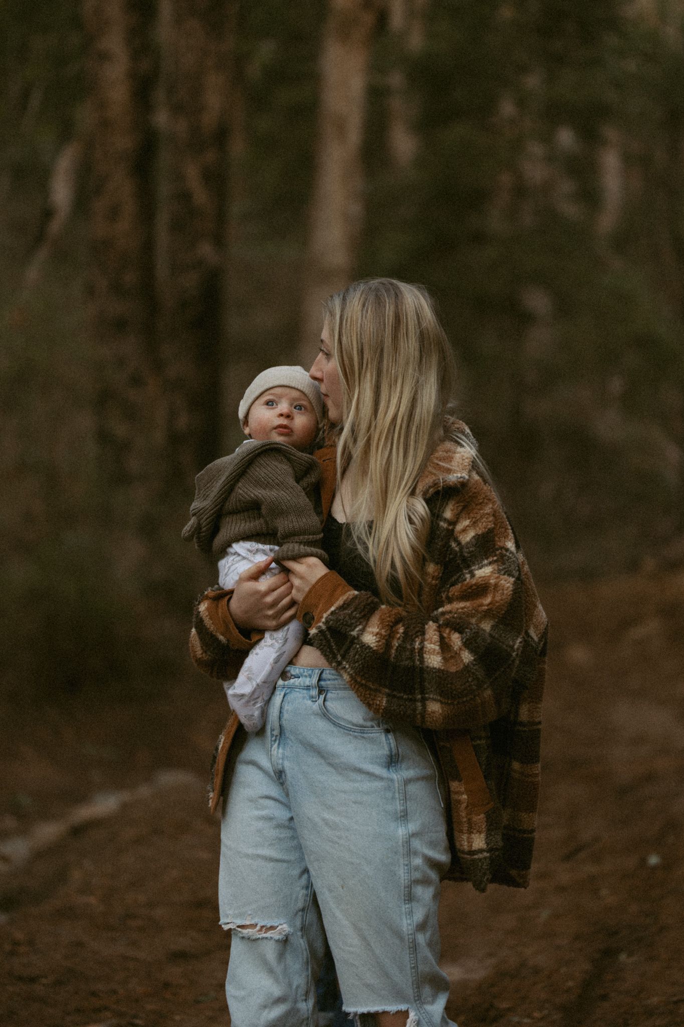 Parent holds child while wearing a cozy plaid coat and ripped jeans in a moody forest setting at dusk.