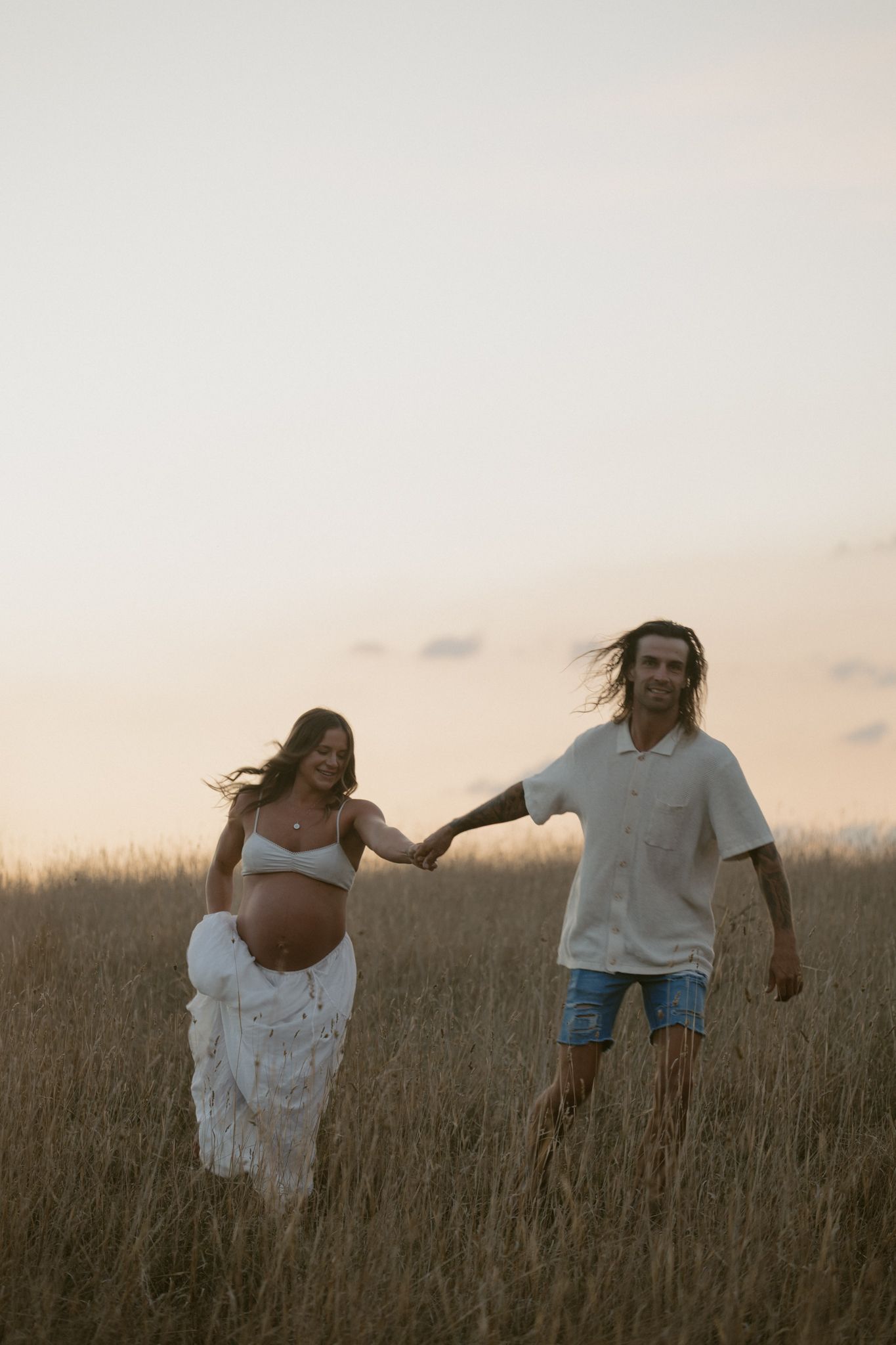 A couple walks hand in hand through a tall grassy field during golden hour sunset.