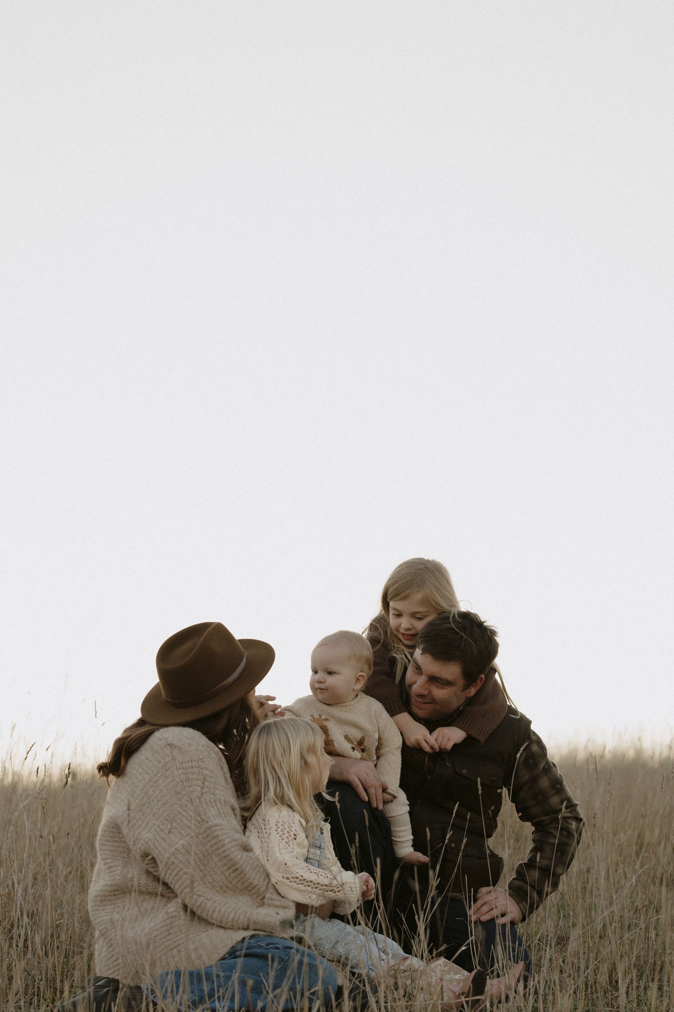 Family of four sits together in tall grass field during golden hour wearing cozy sweaters and casual attire.