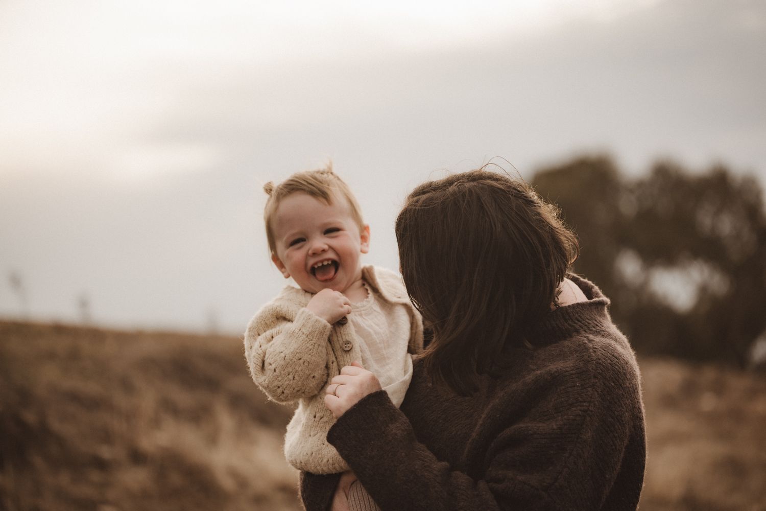 A parent holds a laughing baby in a cozy sweater against an autumn hillside during golden hour.