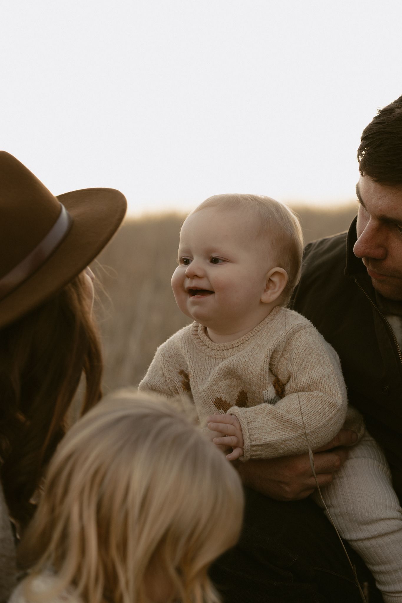 A smiling baby in a cream sweater is held during an intimate outdoor family moment at sunset.