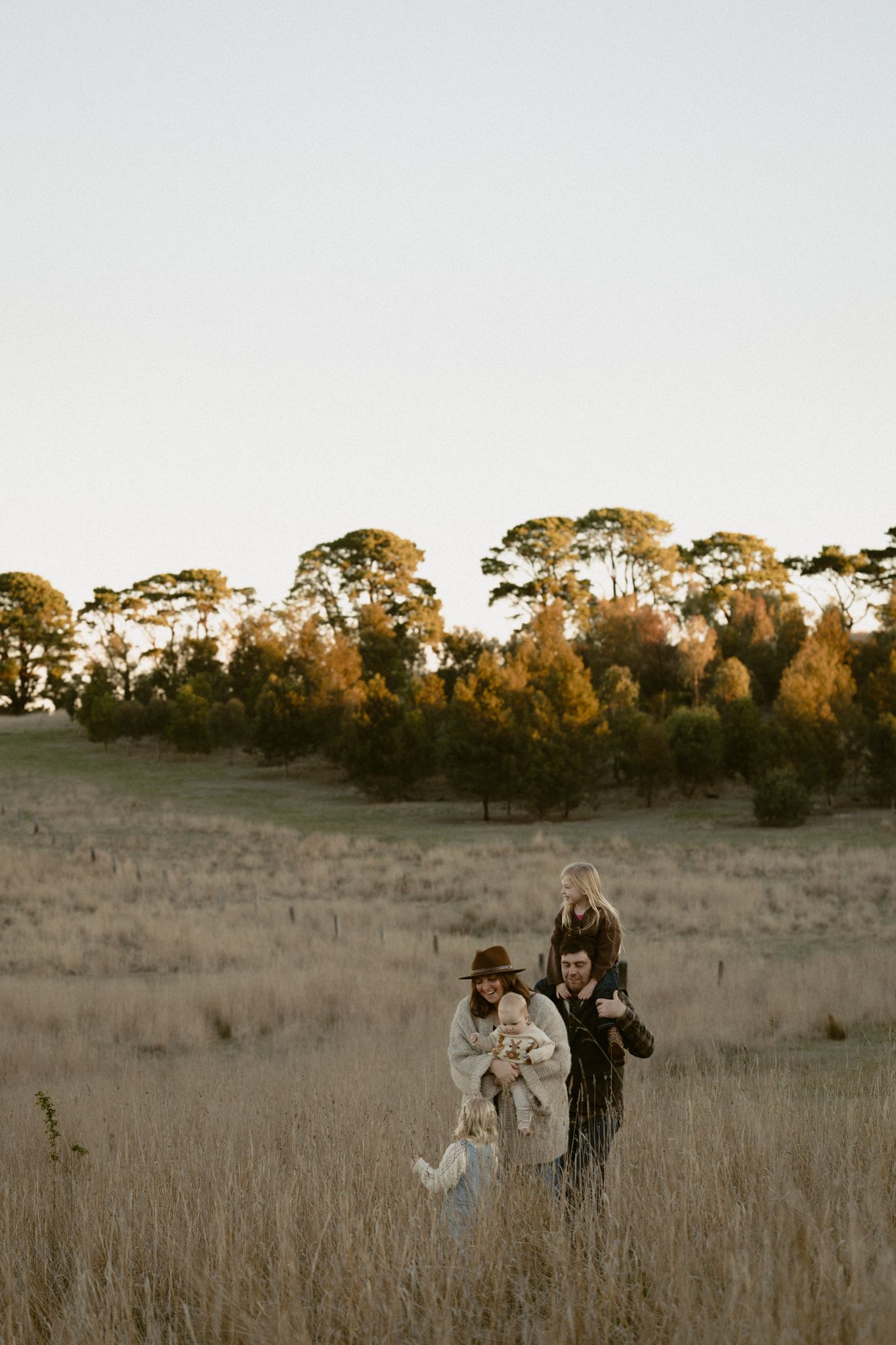 Two people walking through a golden grassy field at sunset with tall trees silhouetted against the sky in autumn.