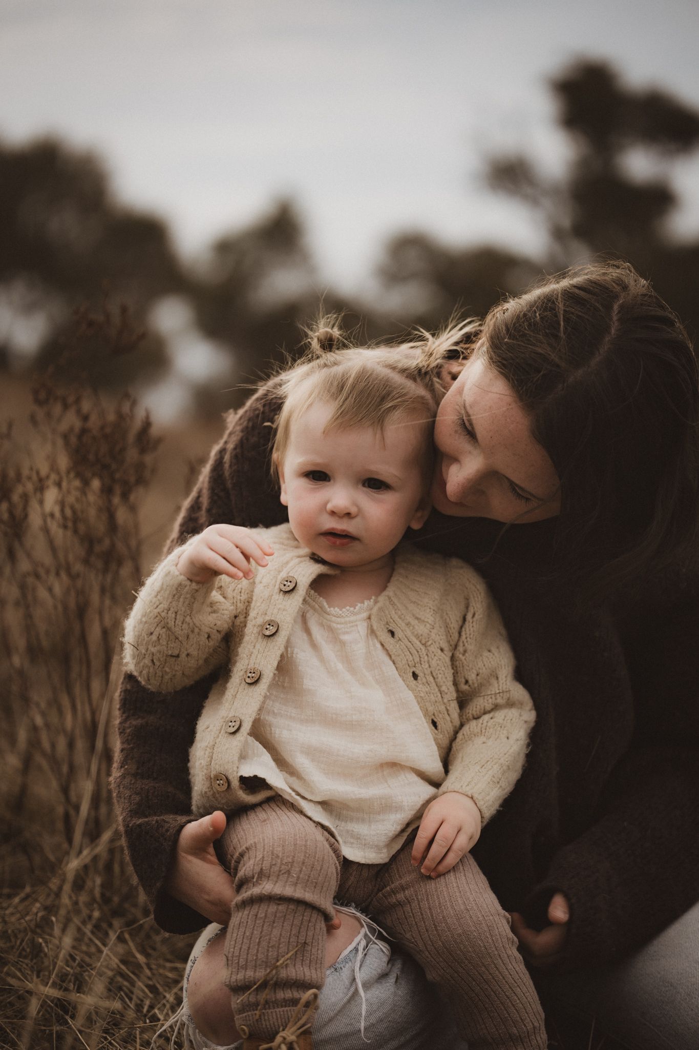 A tender moment captured between parent and child in warm muted tones while sitting among tall dry grass at sunset.