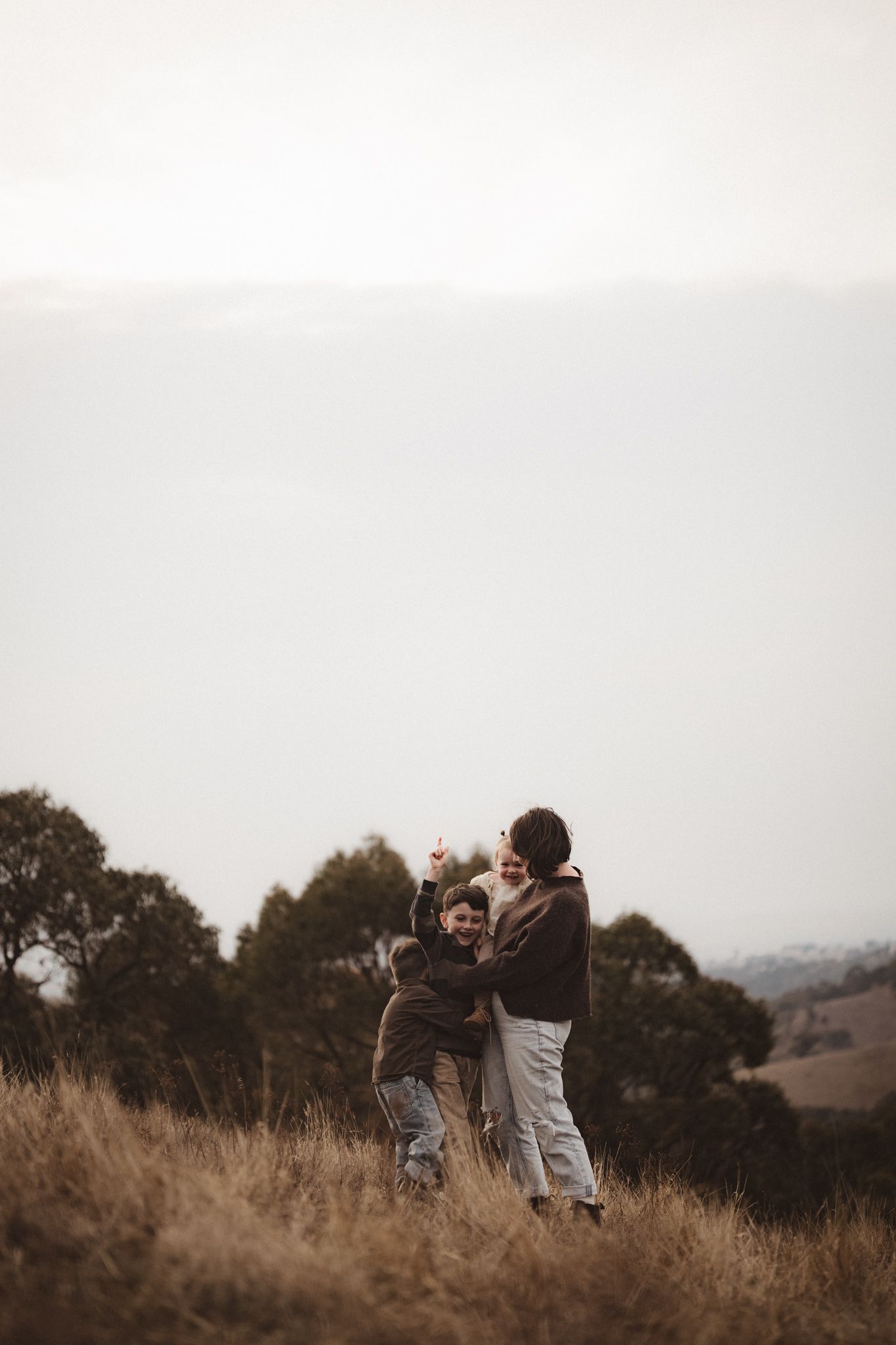 Two people stand together on a hillside with tall golden grass and trees silhouetted against an overcast sky.