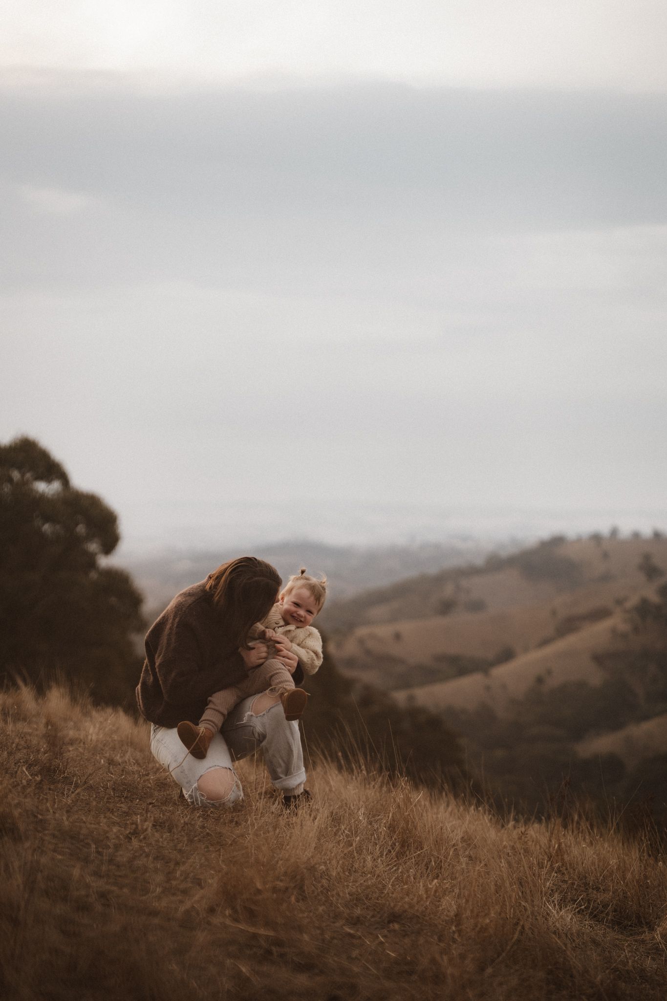 A parent and child share a tender moment on a hilltop during golden hour overlooking rolling hills in the distance.
