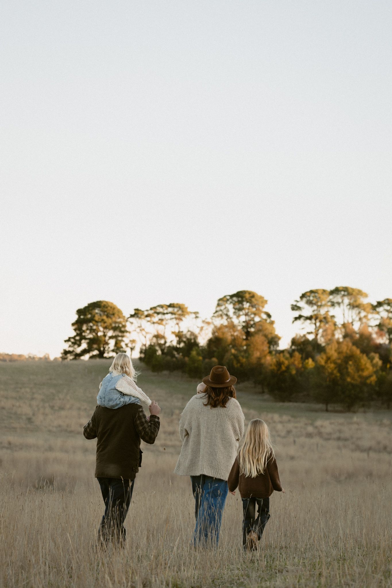 Three people walking together through a golden field at sunset with tall trees silhouetted against the sky in the distance.