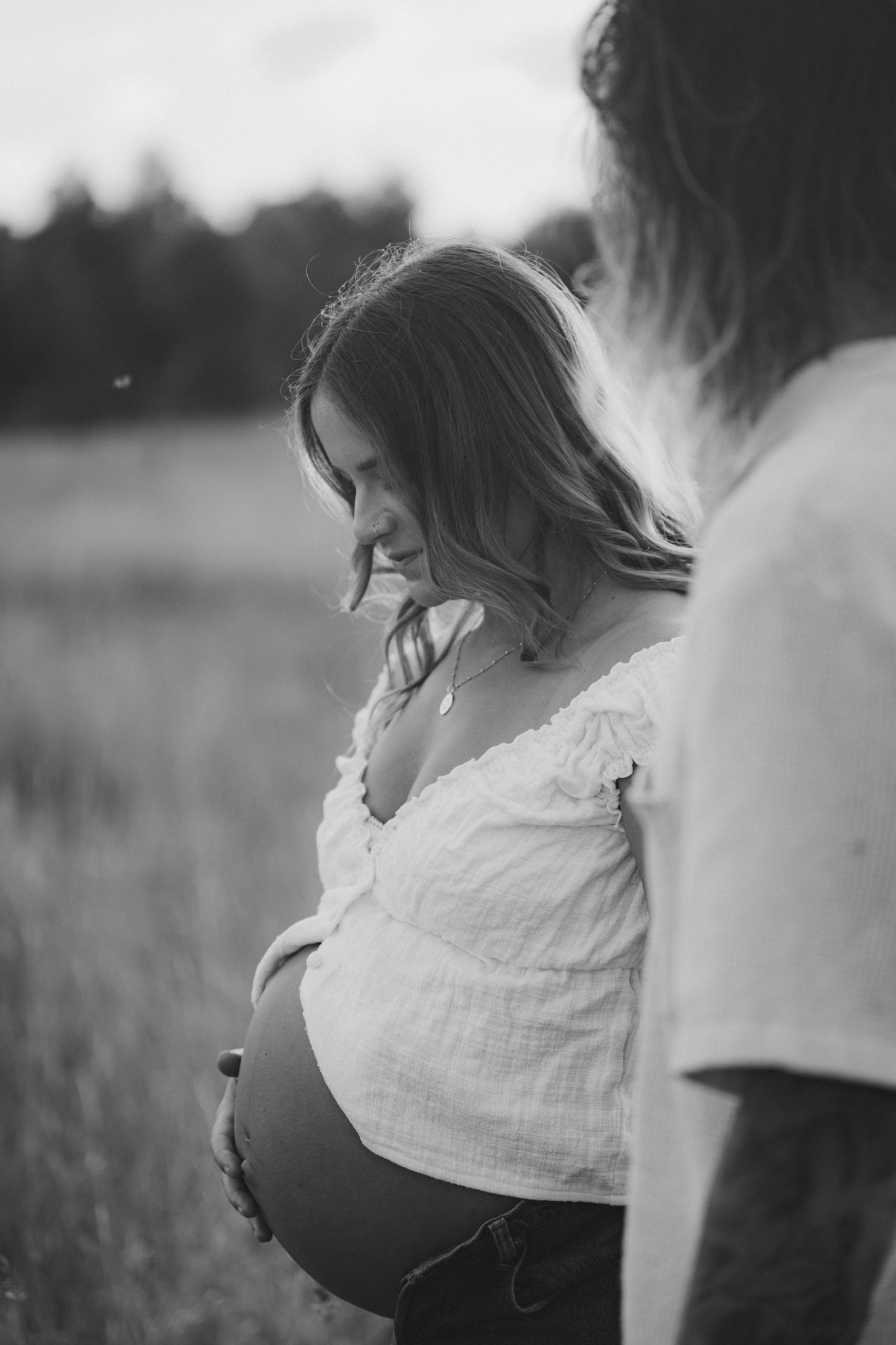 A pregnant woman in a white blouse stands in a field at sunset while being photographed in black and white.