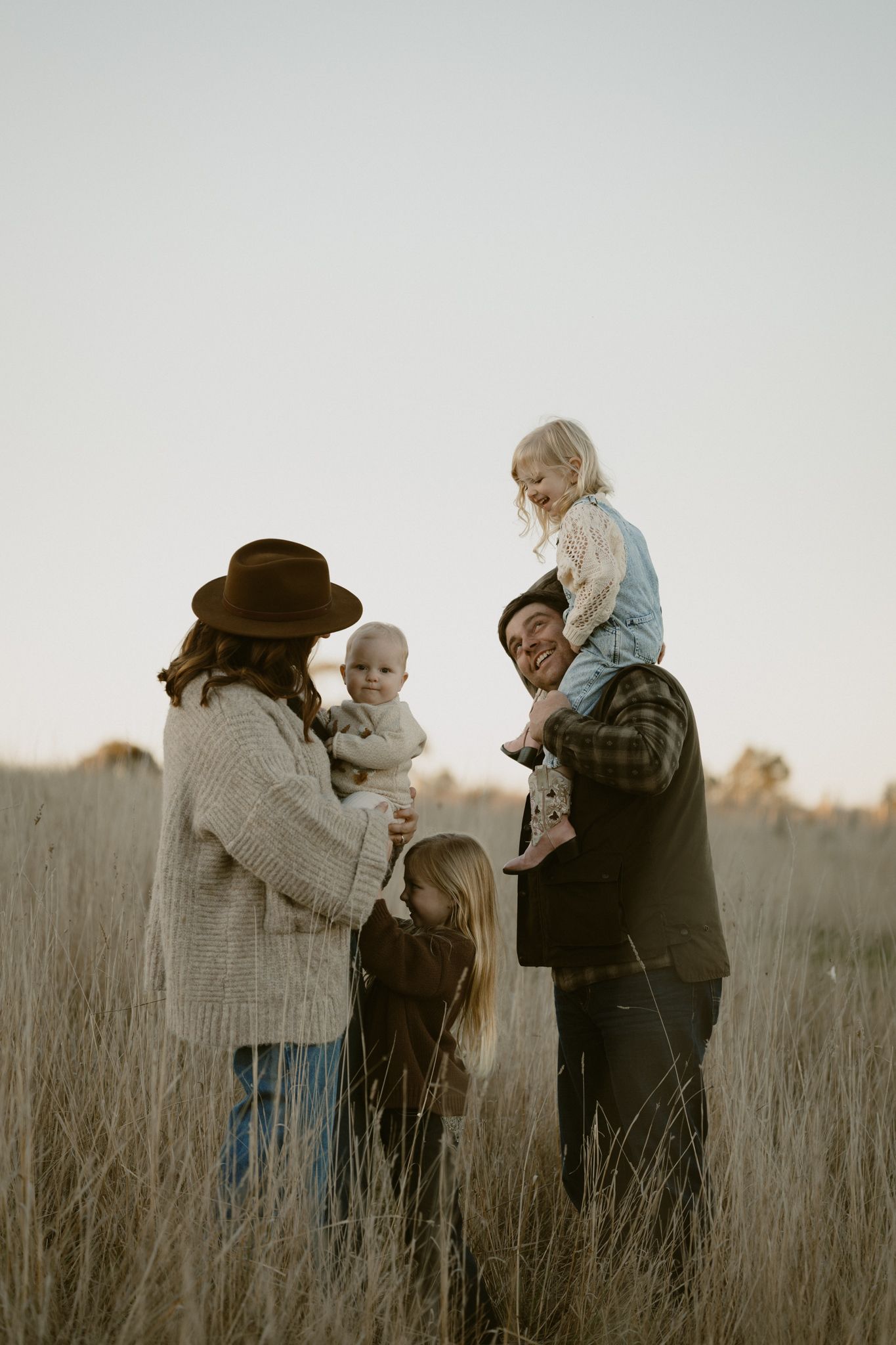 Family stands together in a field of tall grass at sunset enjoying tender moments with a small child.