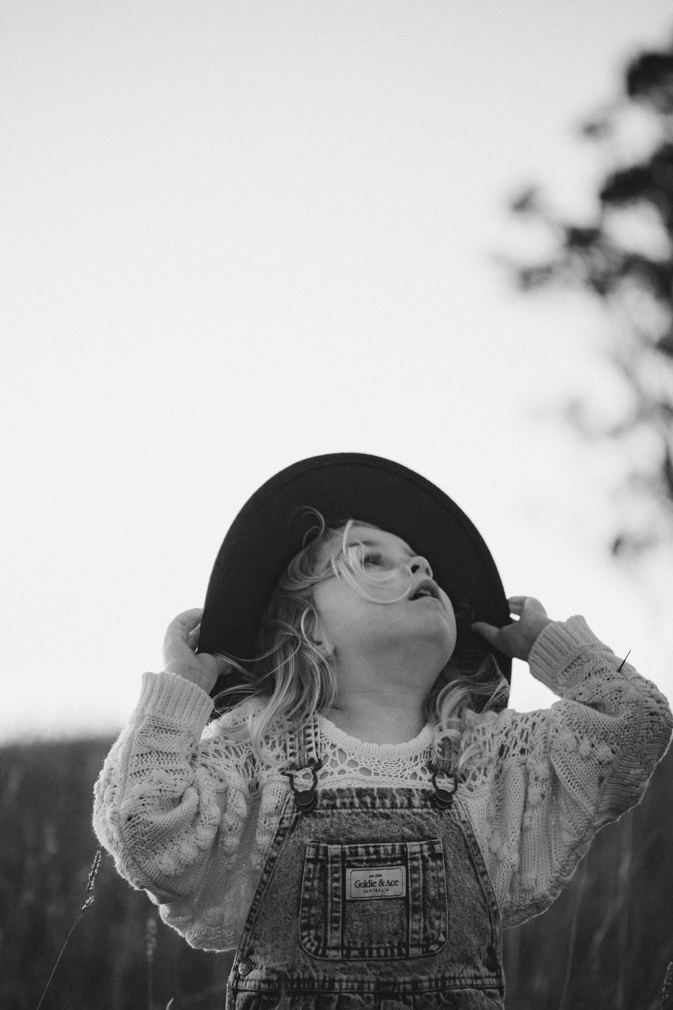 Black and white portrait of a child in overalls and hat looking upward while playing outside.