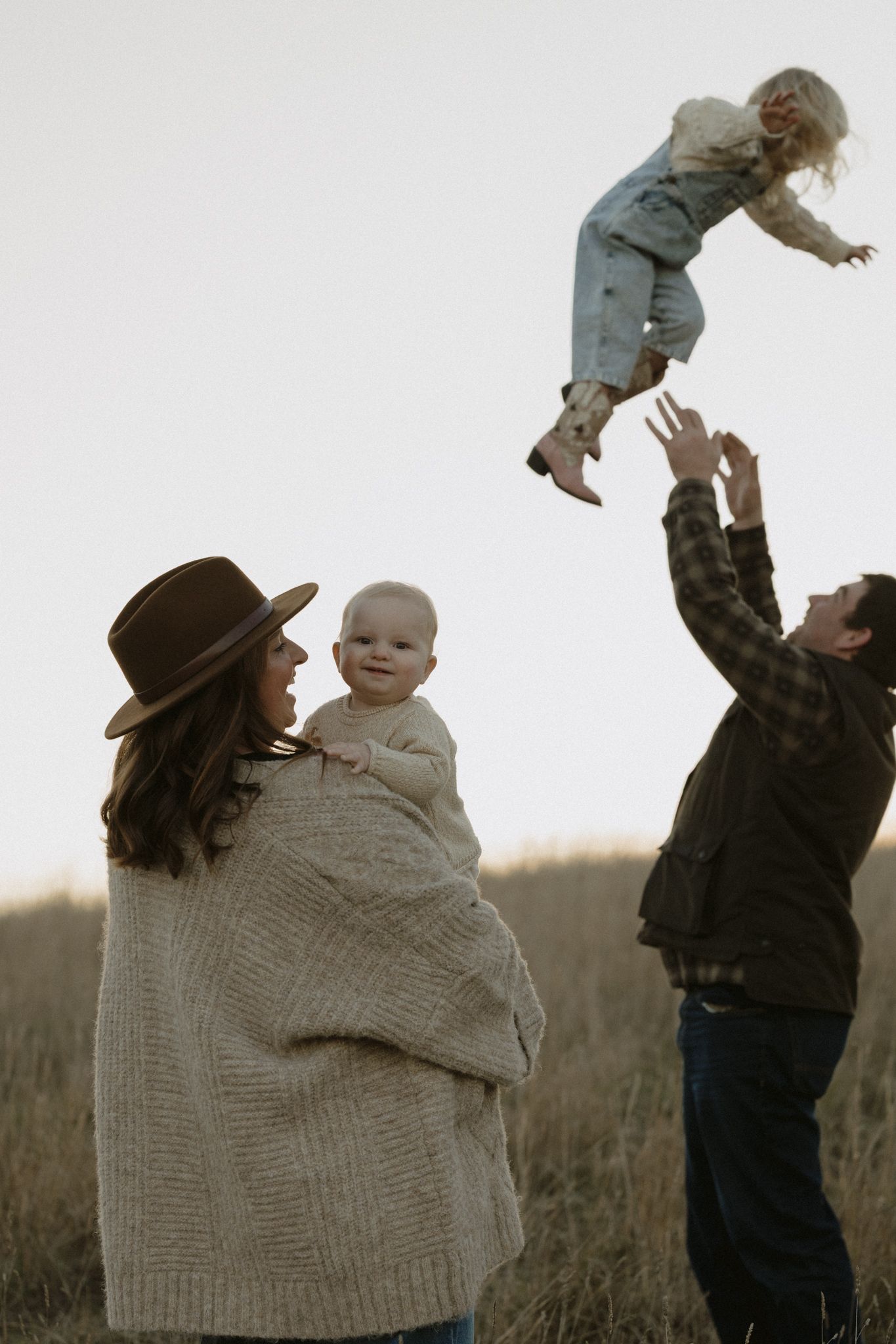 Family enjoying playful moment in field with parent tossing child in air while other parent holds baby in cozy sweater.