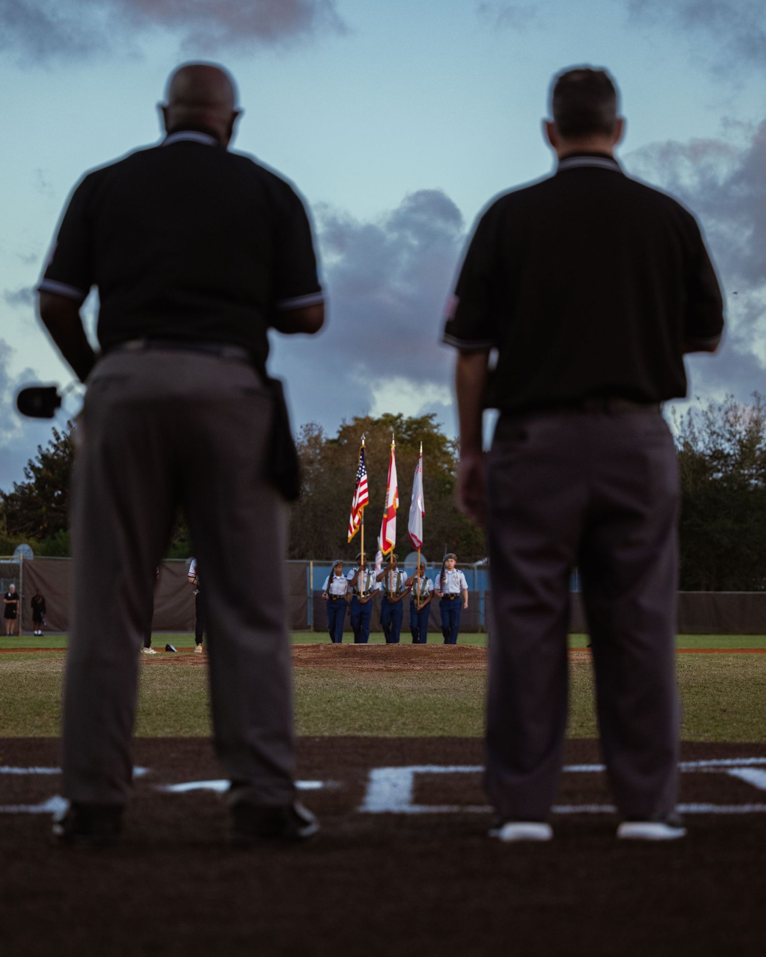 Silhouetted umpires observe flag presentation during sunset at a baseball game, capturing ceremonial moment in South Florida.