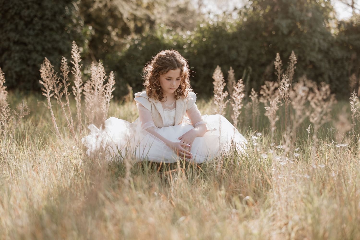 A person in a white dress sits peacefully among tall grasses during golden hour in a dreamy outdoor setting.