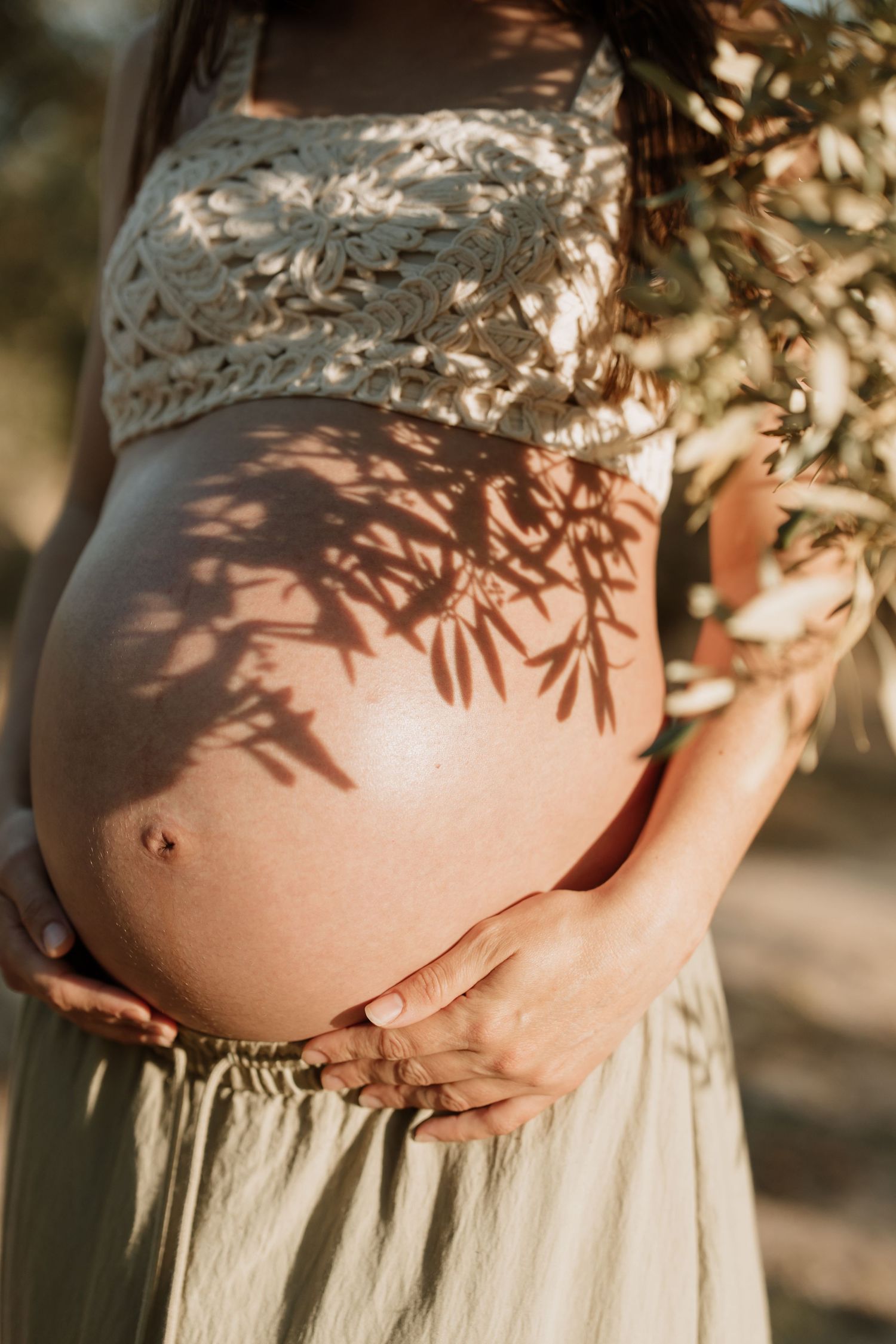Close-up of pregnant belly with lace dress detail and tree shadows in golden sunlight.