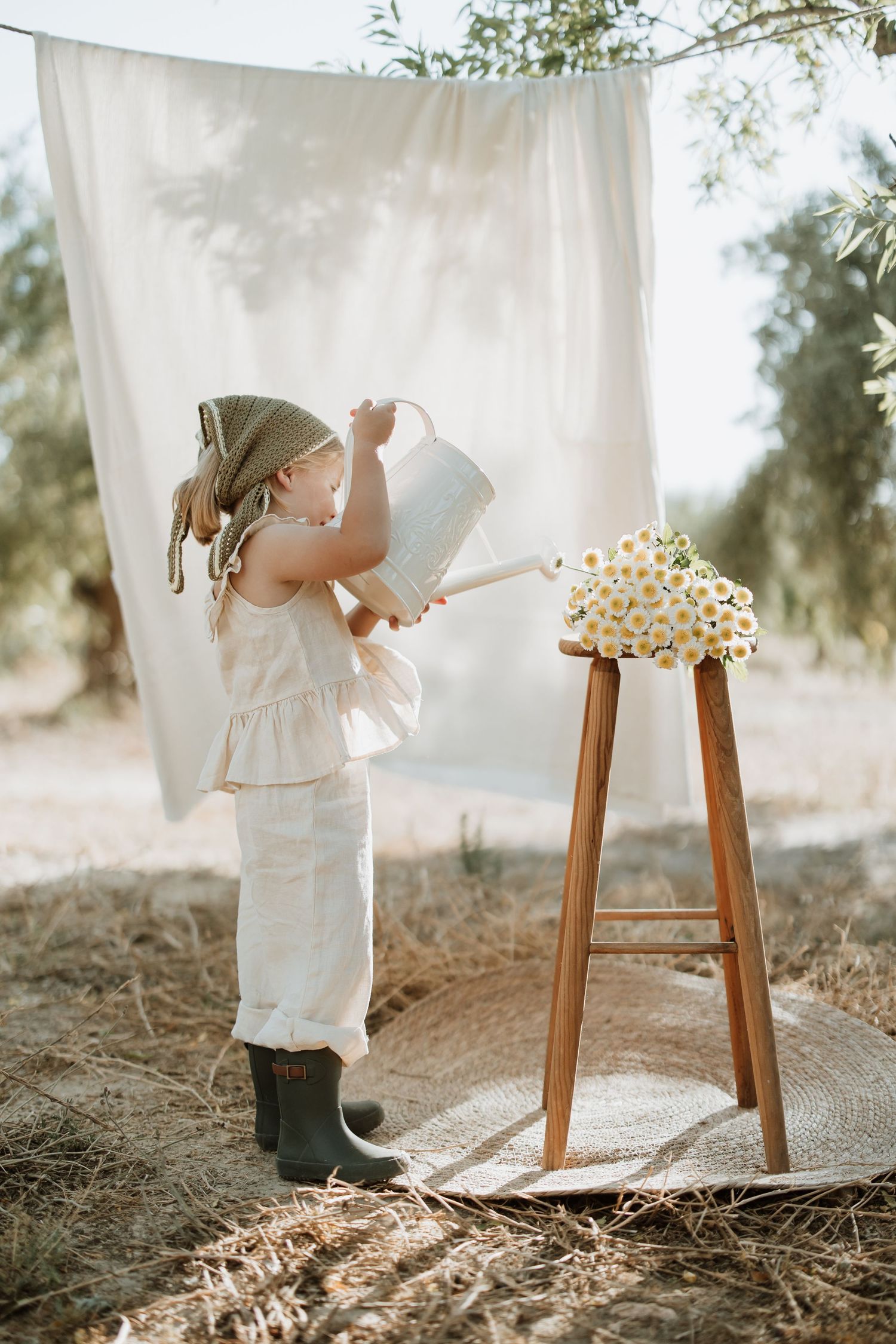 Figure in white dress playing near hanging linens and wooden stool in natural setting.