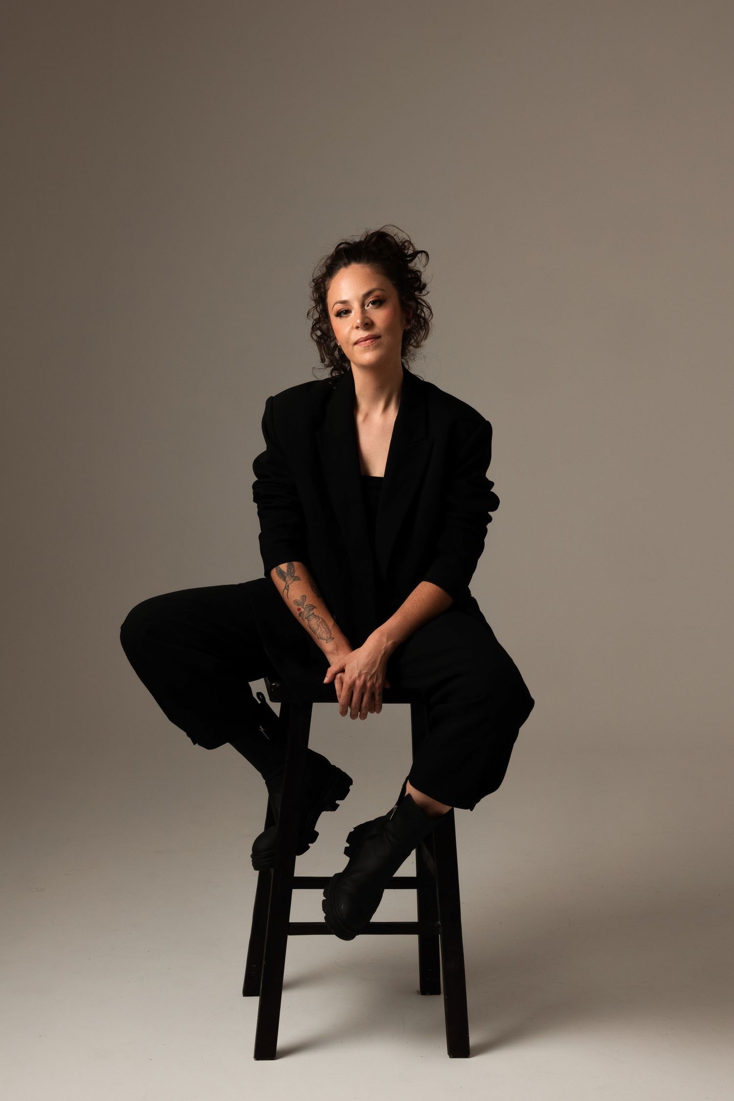 Studio portrait of someone in an all-black outfit seated on a wooden stool against neutral background.