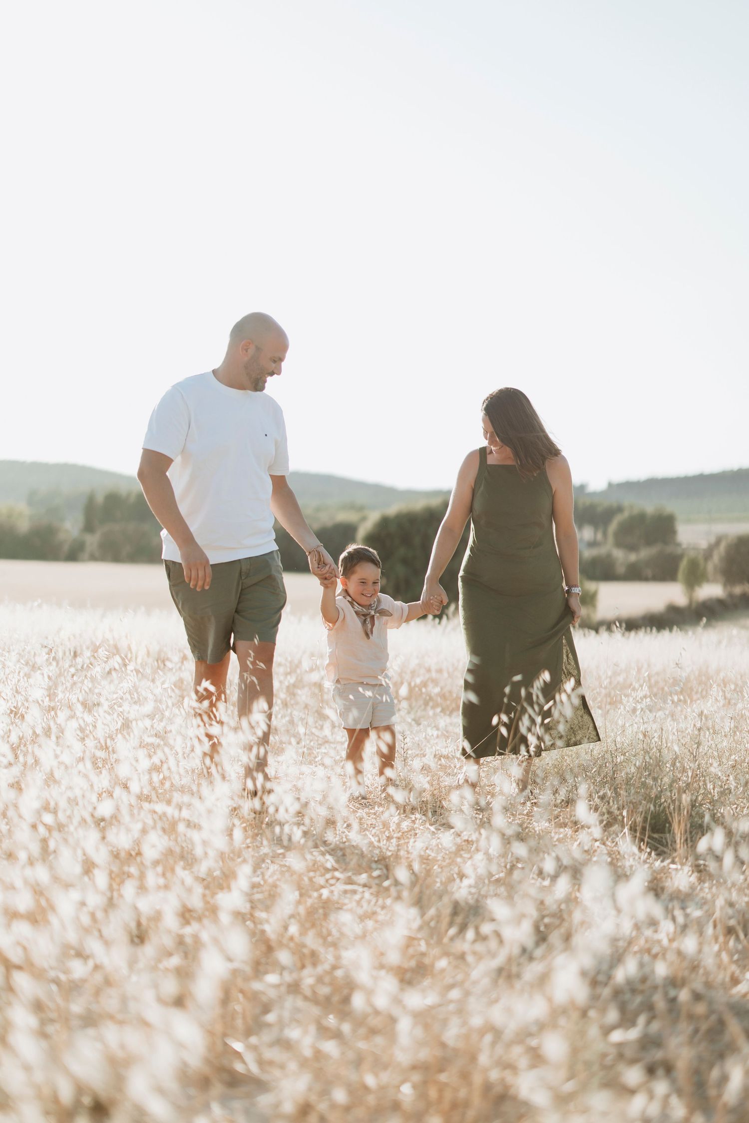 A family walks together through a sunlit meadow of tall grass during golden hour while holding hands.