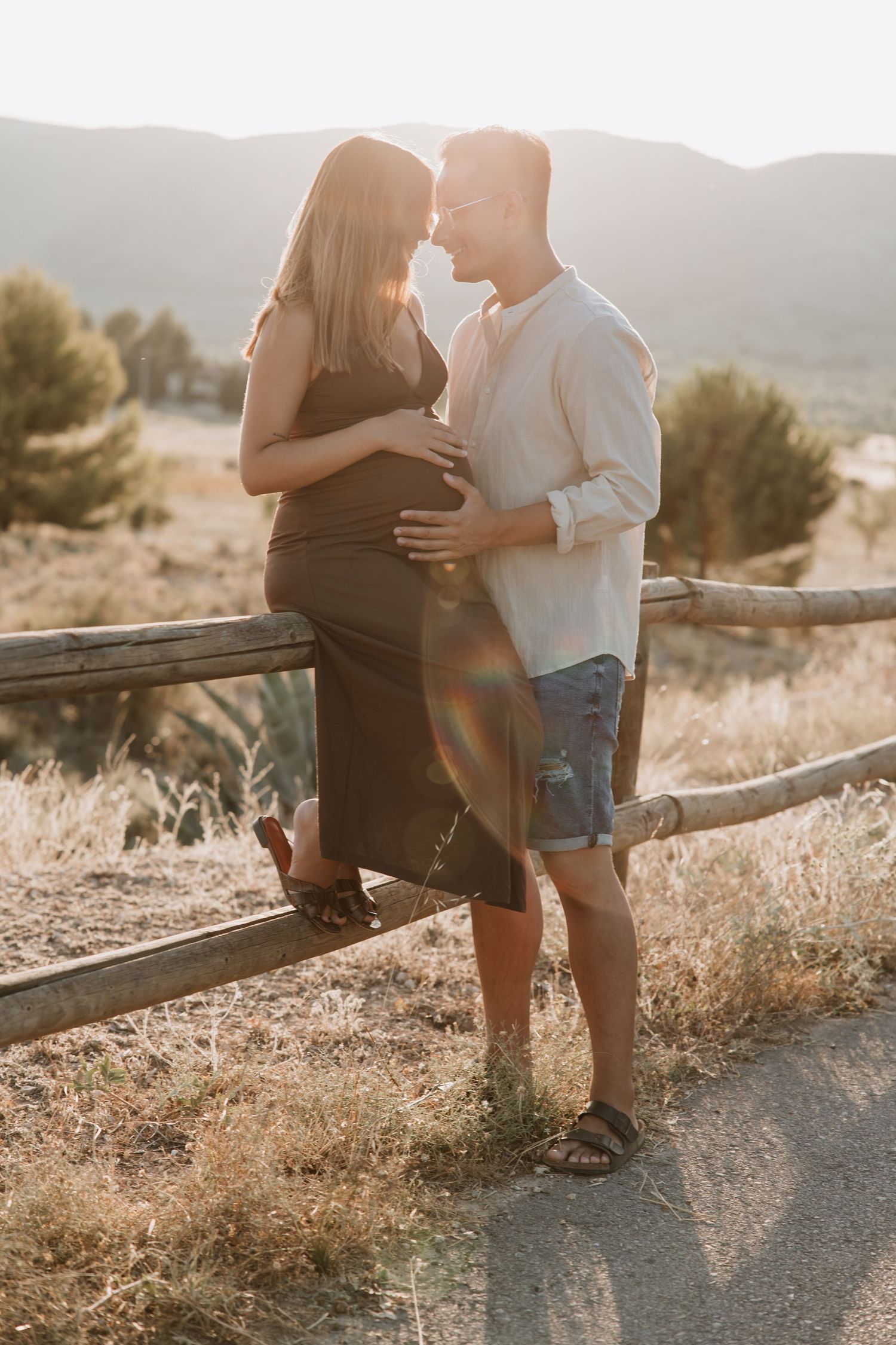 A couple in casual attire embraces at sunset on a rustic wooden fence with mountains and golden light in the background.