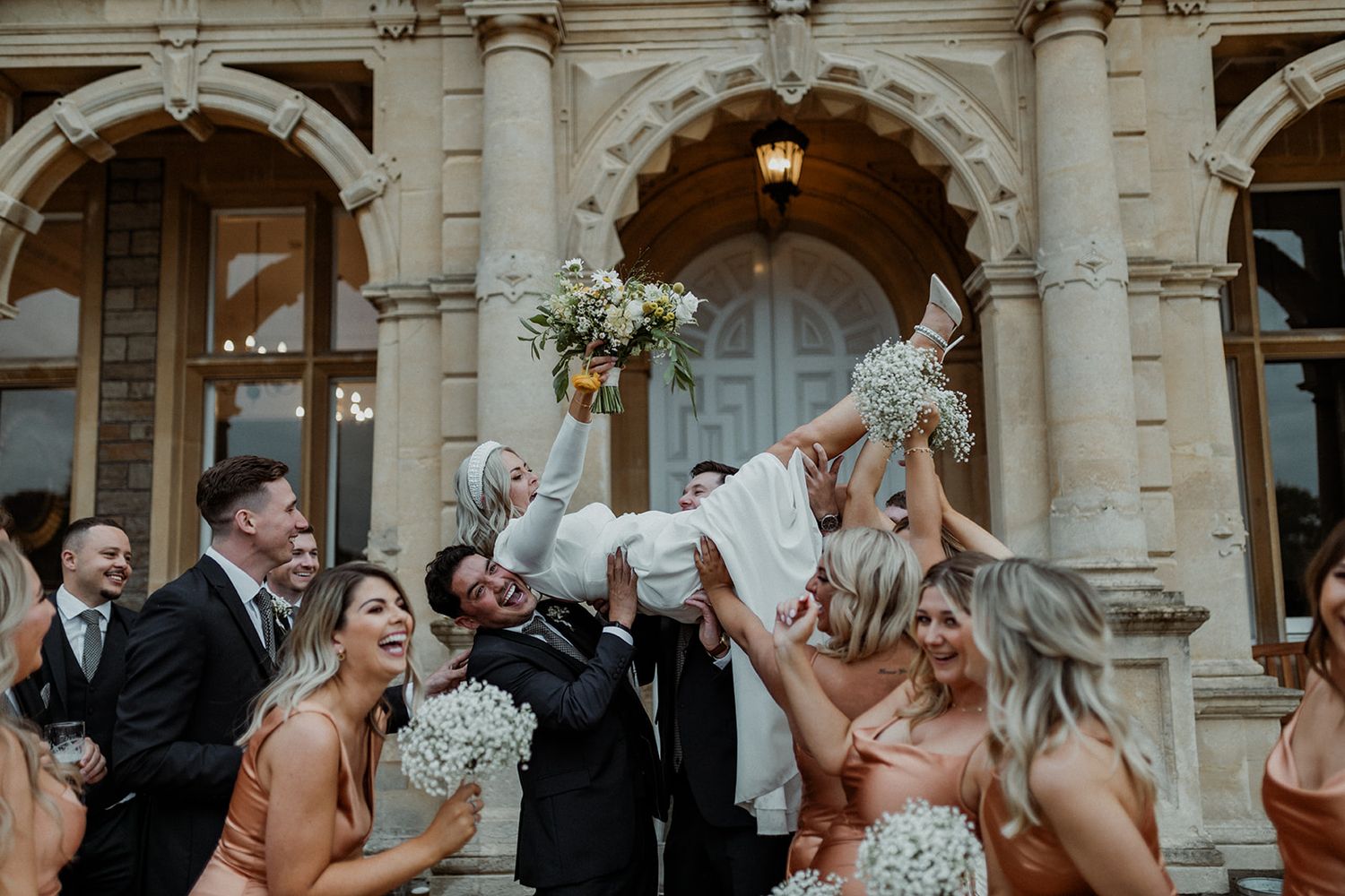 A panoramic shot of a wedding party cheering and celebrating outside an ornate stone building with classical architecture.