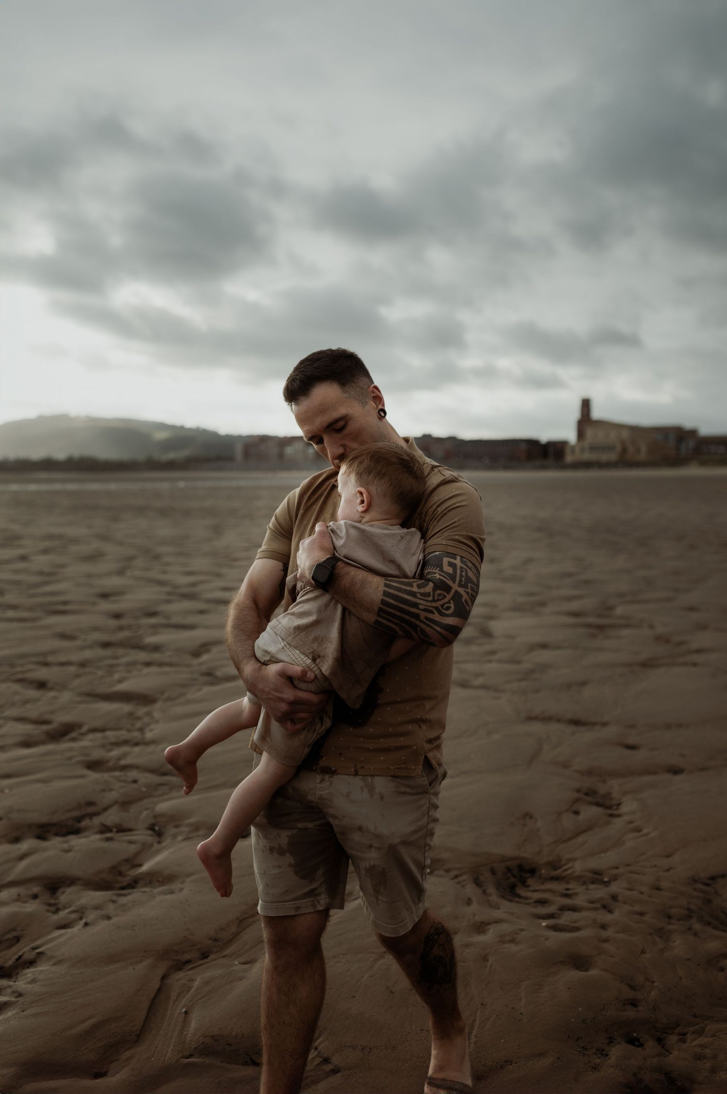 A parent holding a small baby stands on a moody beach at dusk with a distant lighthouse in the background.