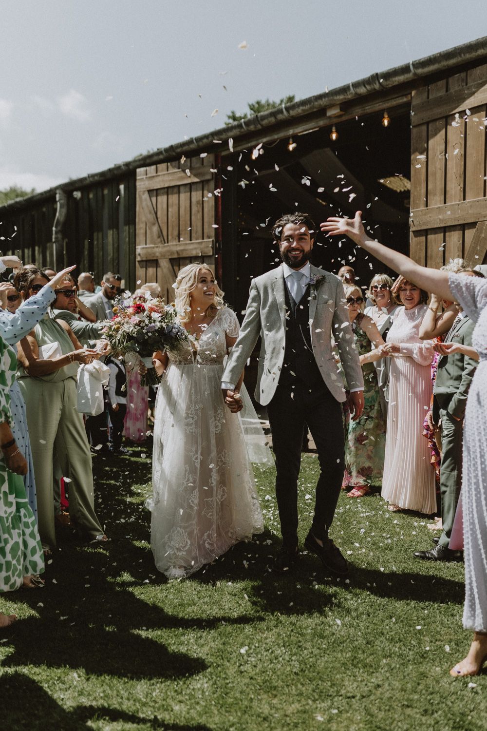 Wedding party celebrating outside with guests tossing petals and confetti on a sunny day.