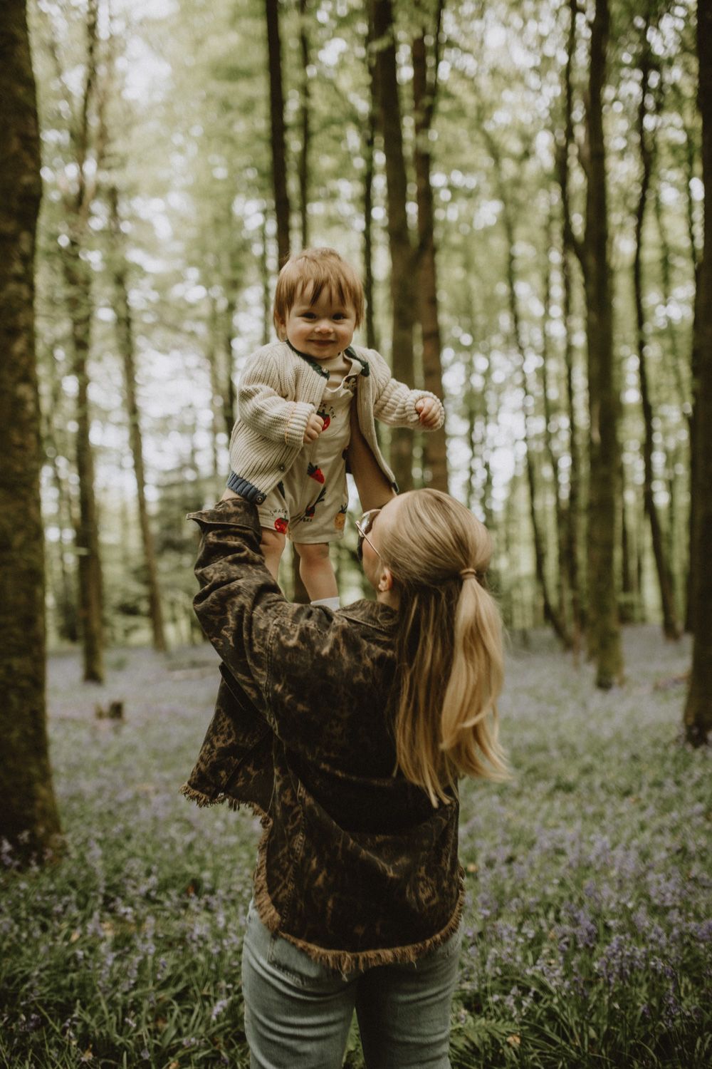 A tender moment captured in a forest clearing with wildflowers as someone lifts a small child wearing a light sweater.