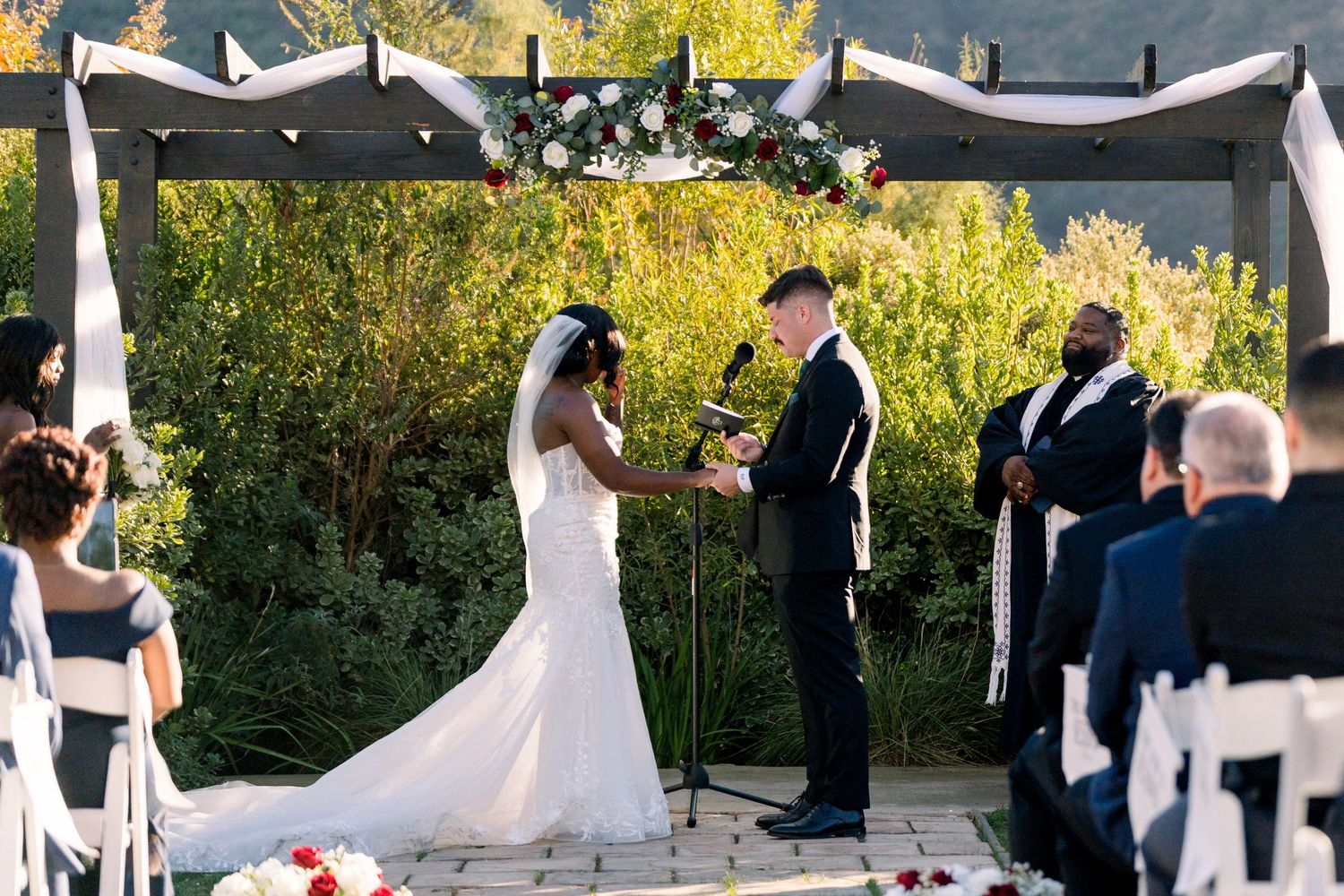 Outdoor wedding ceremony under white arbor with couple exchanging vows surrounded by guests.