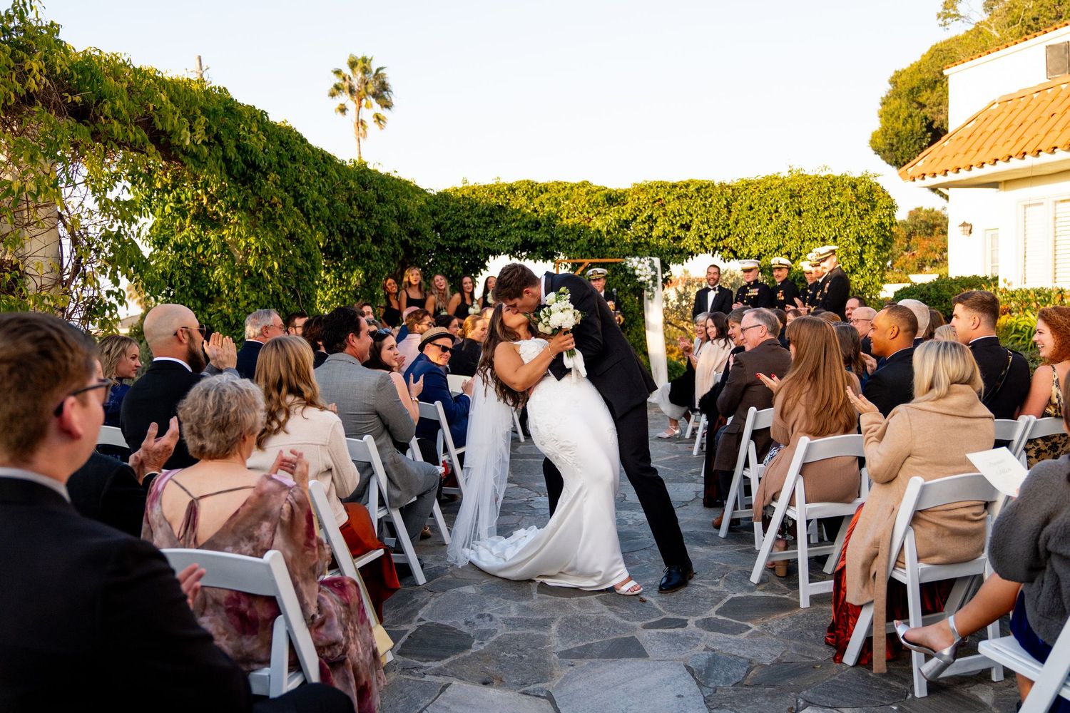 Wedding ceremony taking place on a stone patio surrounded by greenery with guests seated in white chairs.