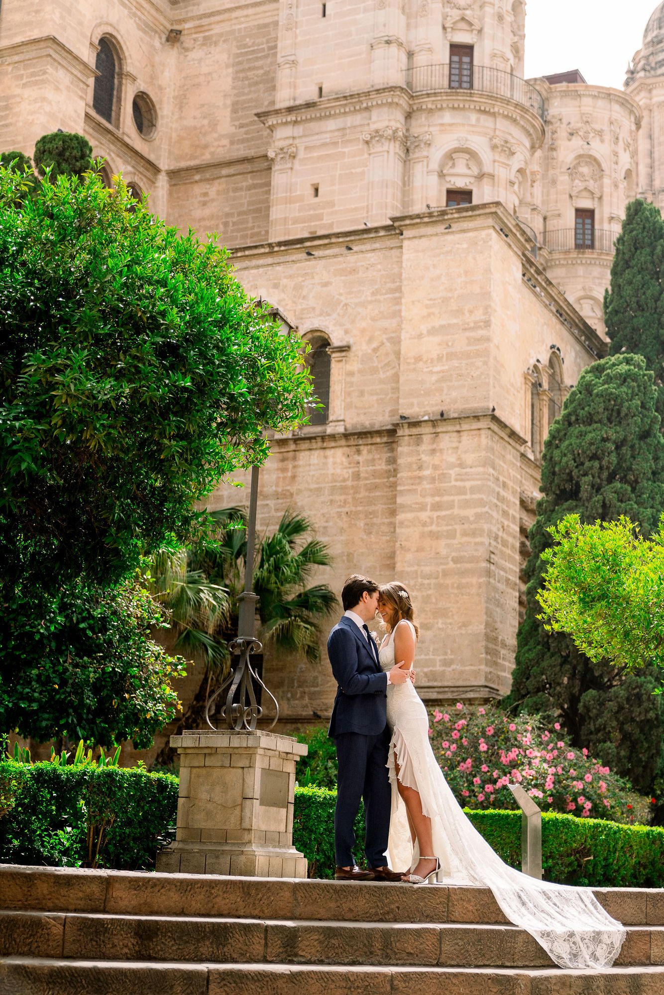A couple shares a romantic moment on stone steps outside a grand historic cathedral with lush greenery and gardens.